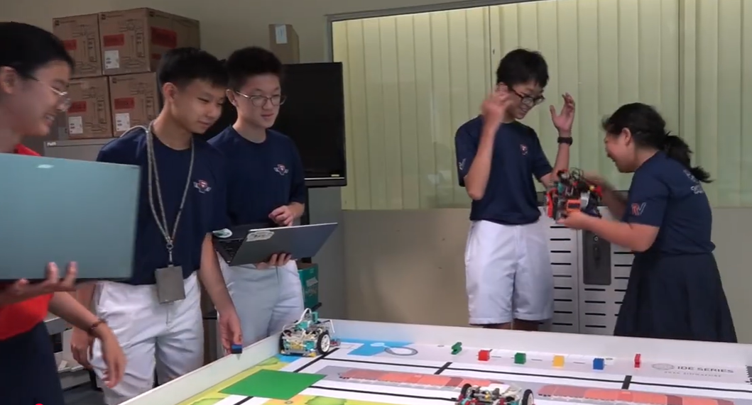Five students in navy uniforms stand around a colorful robotics competition table with small robots and colored blocks.