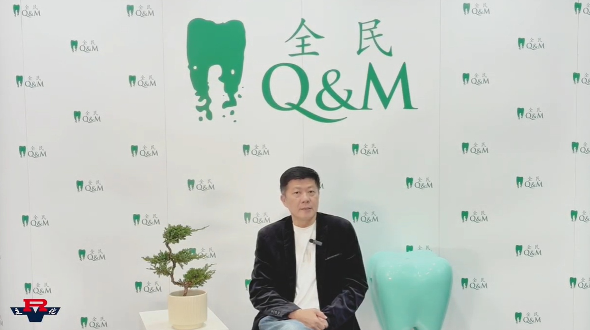 Man in black jacket sits before a Q&M dental branded backdrop, with a small bonsai and tooth-shaped decor nearby.