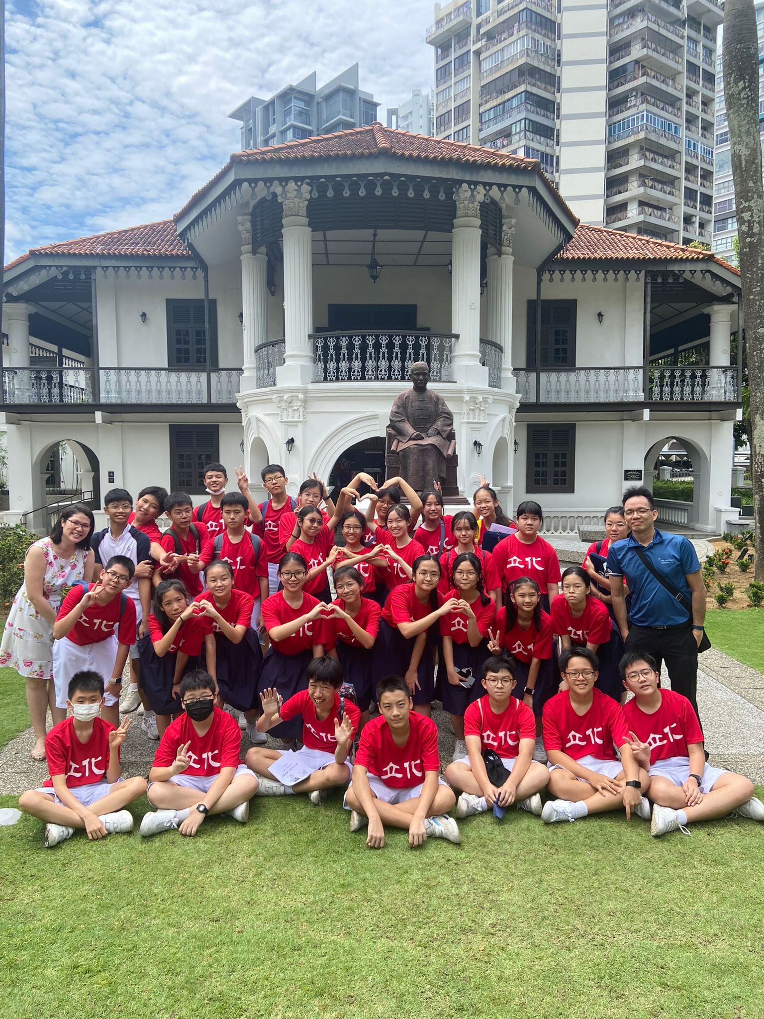 A large group of students in red shirts pose on grass in front of a white colonial building with a bronze statue.