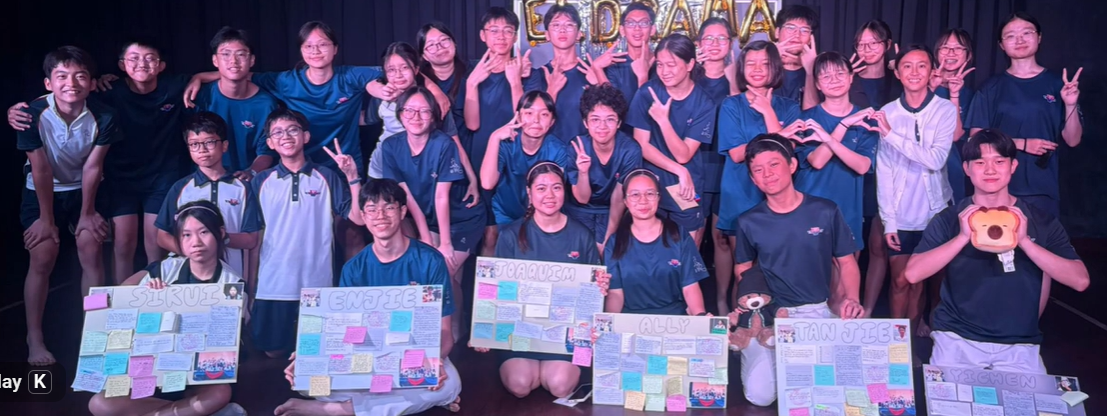 A large group of students in blue uniforms pose together, holding colorful handmade poster boards with names and messages.