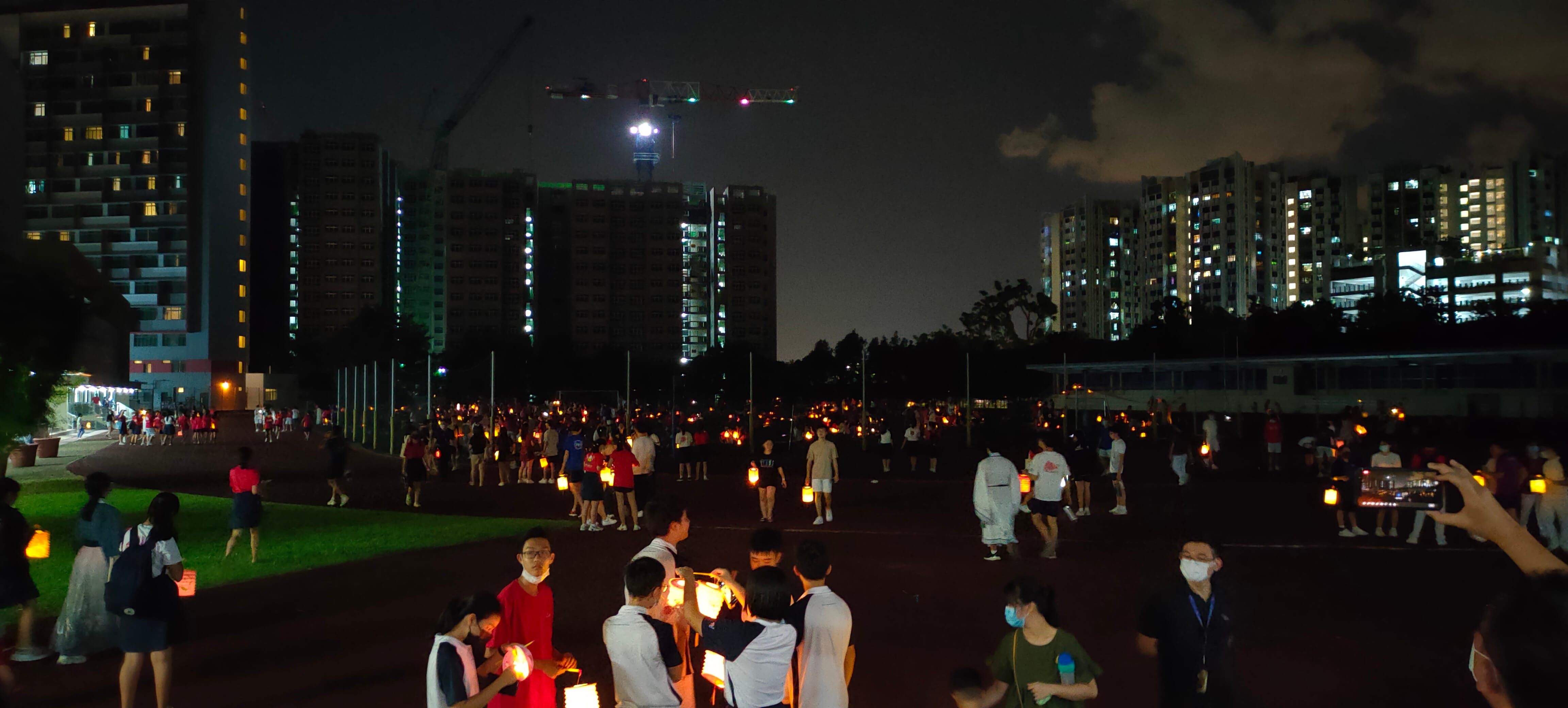A large crowd of people holding glowing lanterns on an outdoor field at night with tall lit buildings in the background.