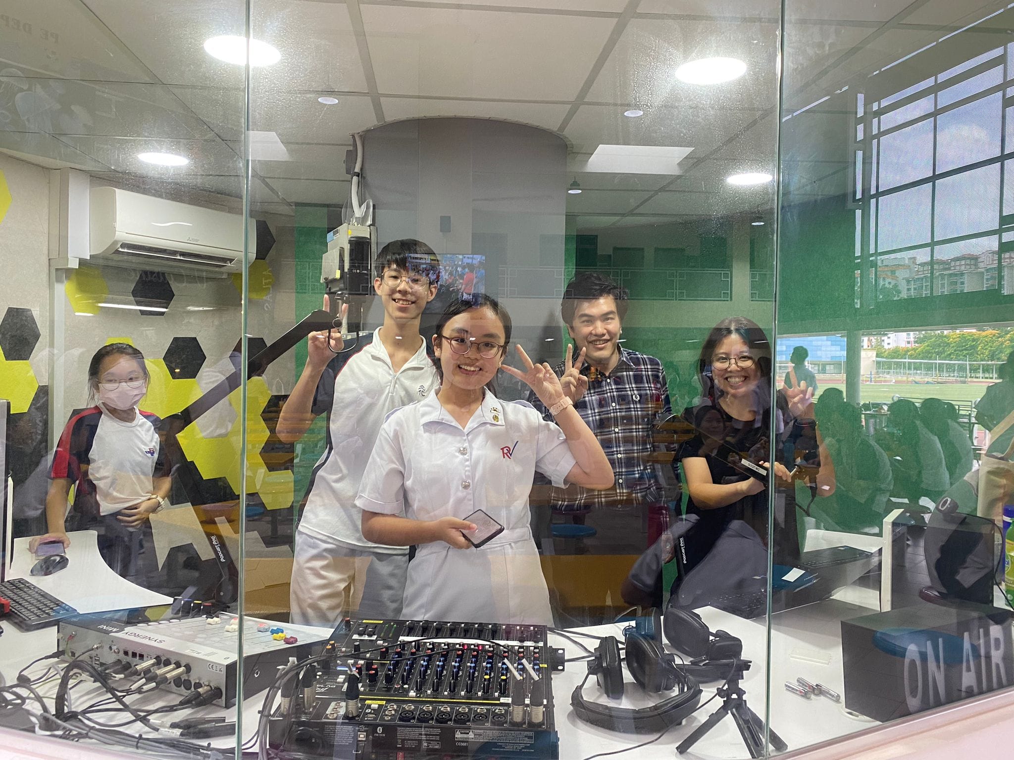 Five people smile and pose in a school radio studio with an audio mixer, headphones, and an On Air sign.