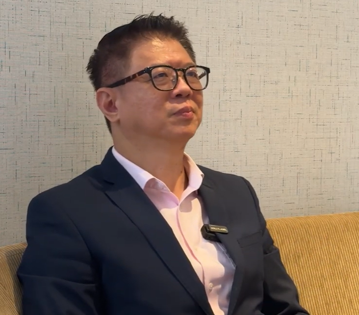 A middle-aged man wearing glasses and a dark blazer over a pink shirt sits on a beige sofa, looking to the side.
