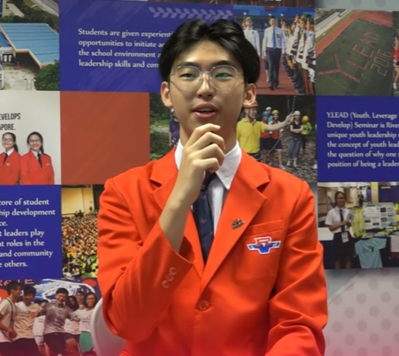 A young male student in a red blazer and tie, wearing glasses, speaking in front of a school display board.