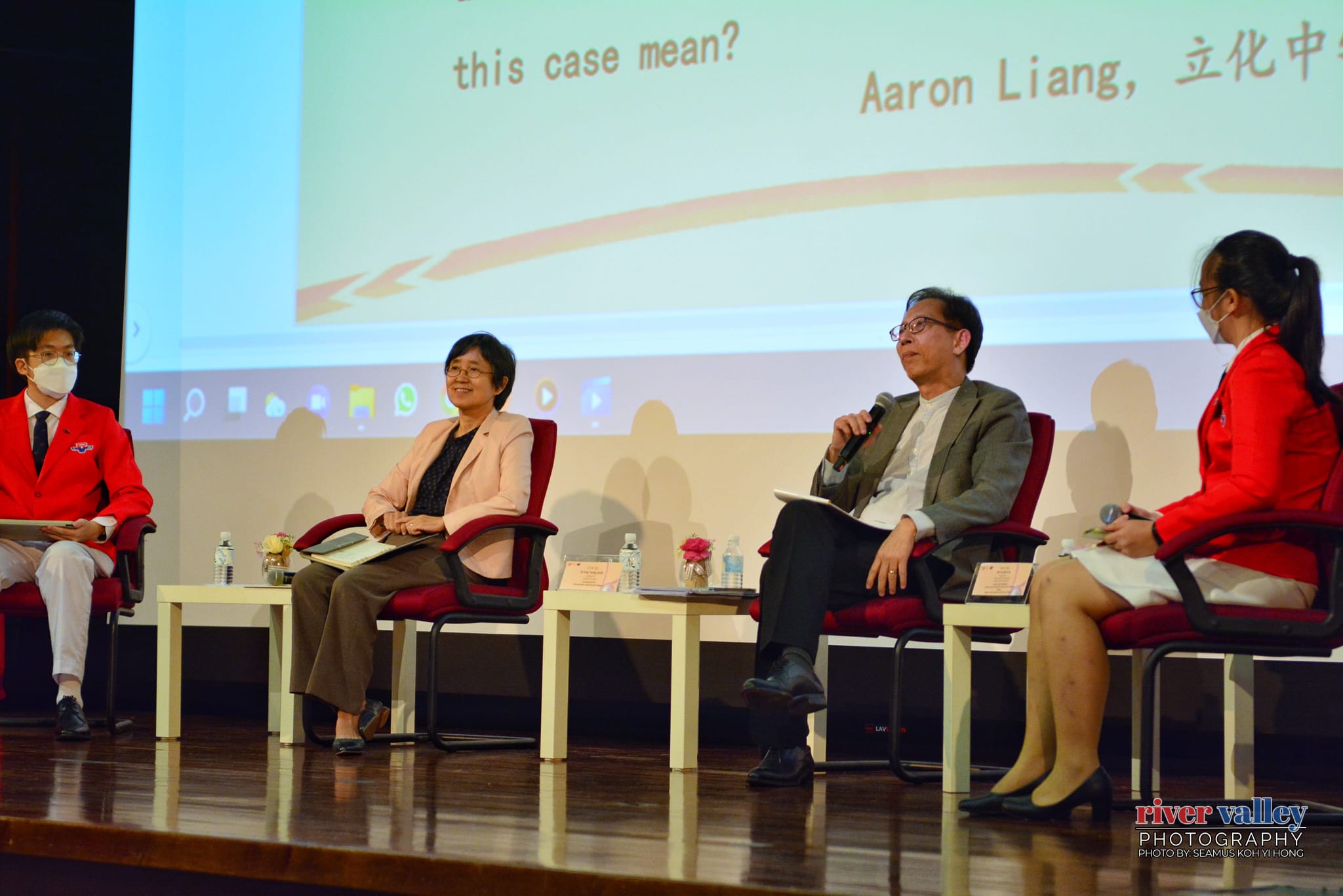 Four people sit on stage in red chairs during a panel discussion, with a projected presentation visible behind them.