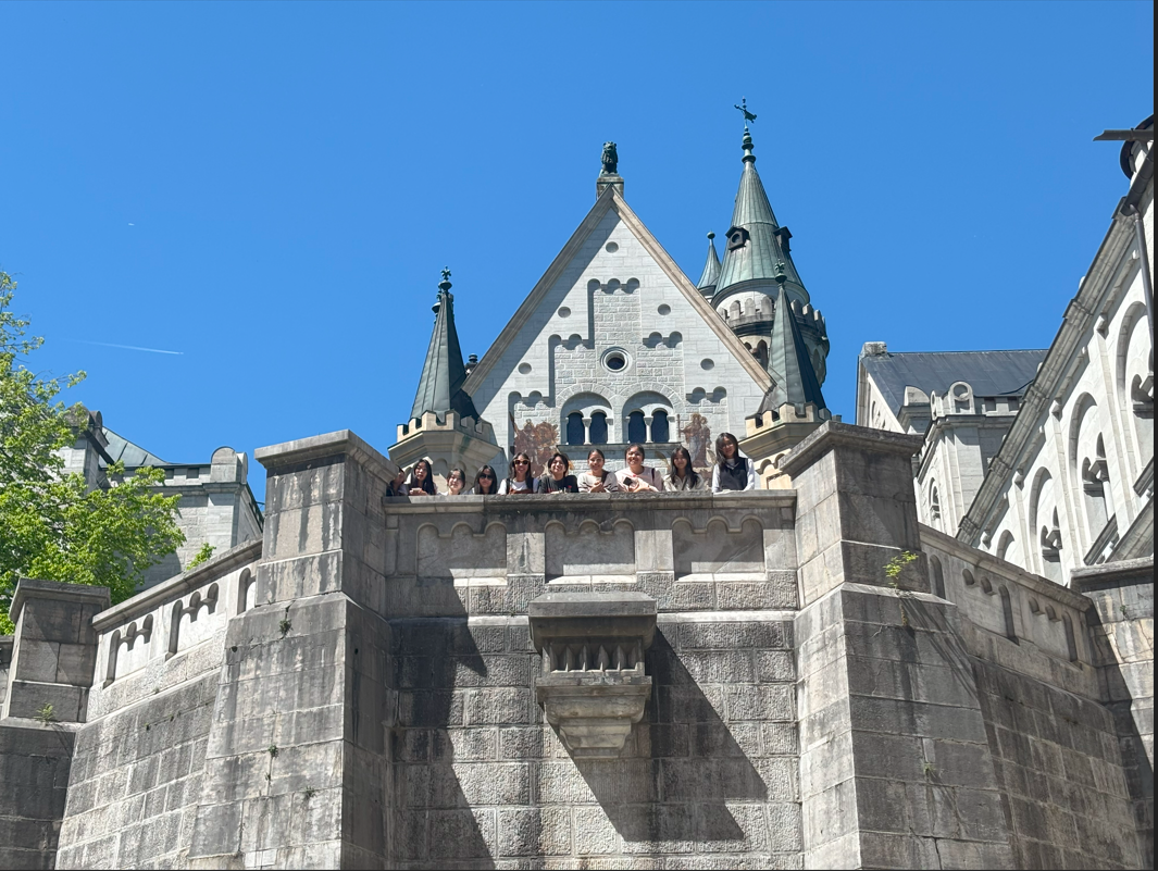 A group of people pose on a balcony of a large stone castle with green spired towers under a clear blue sky.