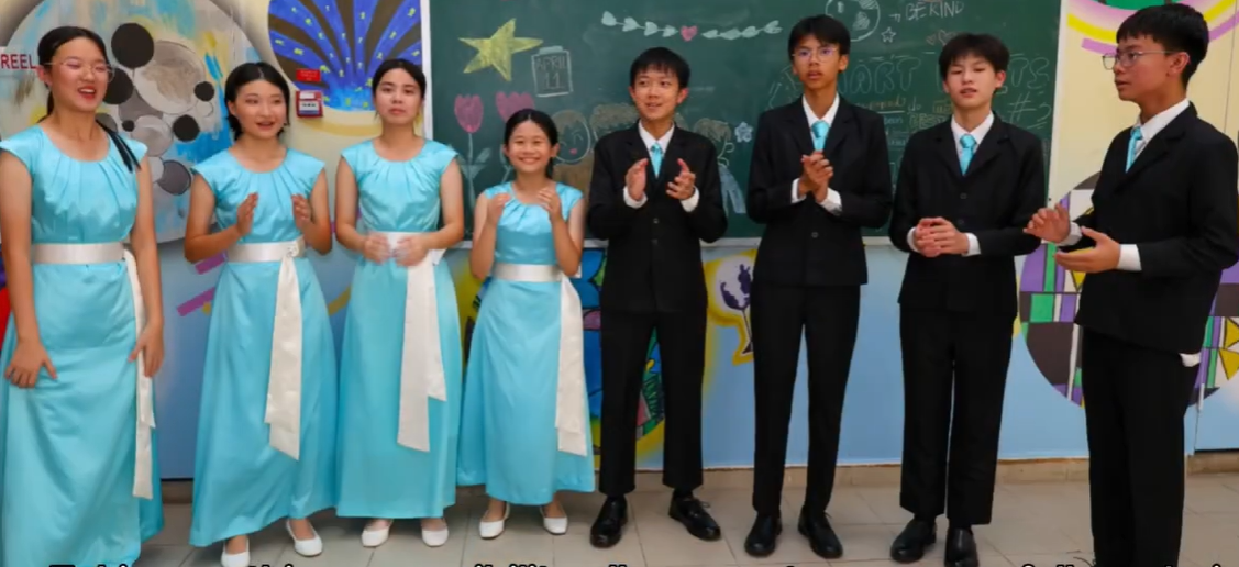 Eight students stand in a classroom, girls in light blue dresses and boys in black suits with teal ties.