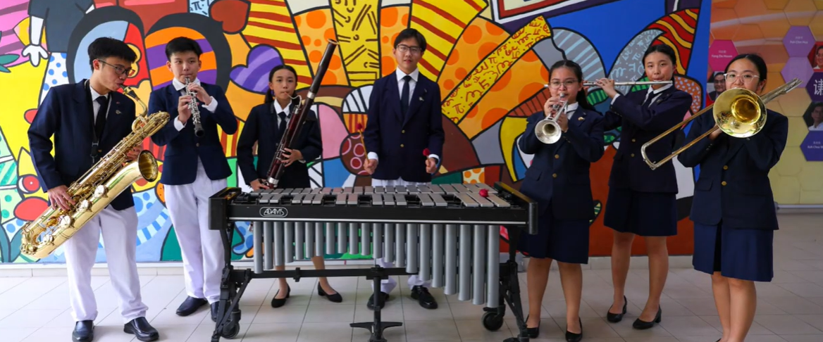 Seven students in school uniforms playing instruments including saxophone, clarinet, bassoon, marimba, trumpet, flute, and trombone before a colorful mural.