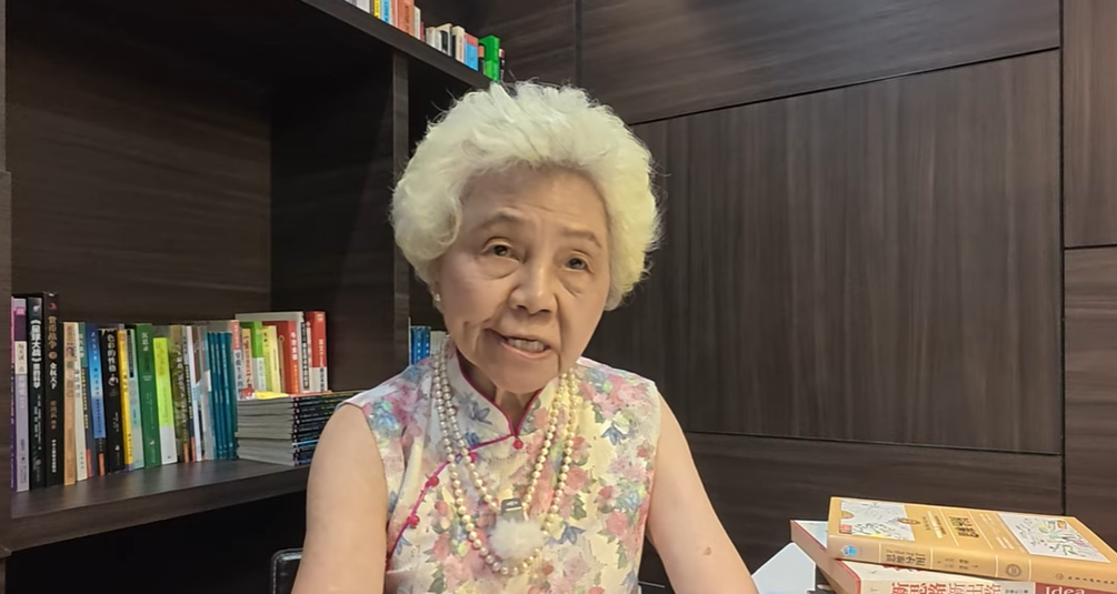 Elderly Asian woman with white hair wearing a floral dress and pearl necklace, seated in front of a bookshelf.