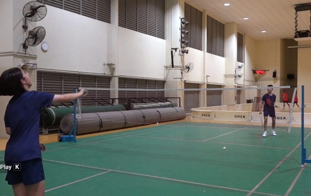 Two players playing badminton on an indoor green court with a net between them and fans on the walls.