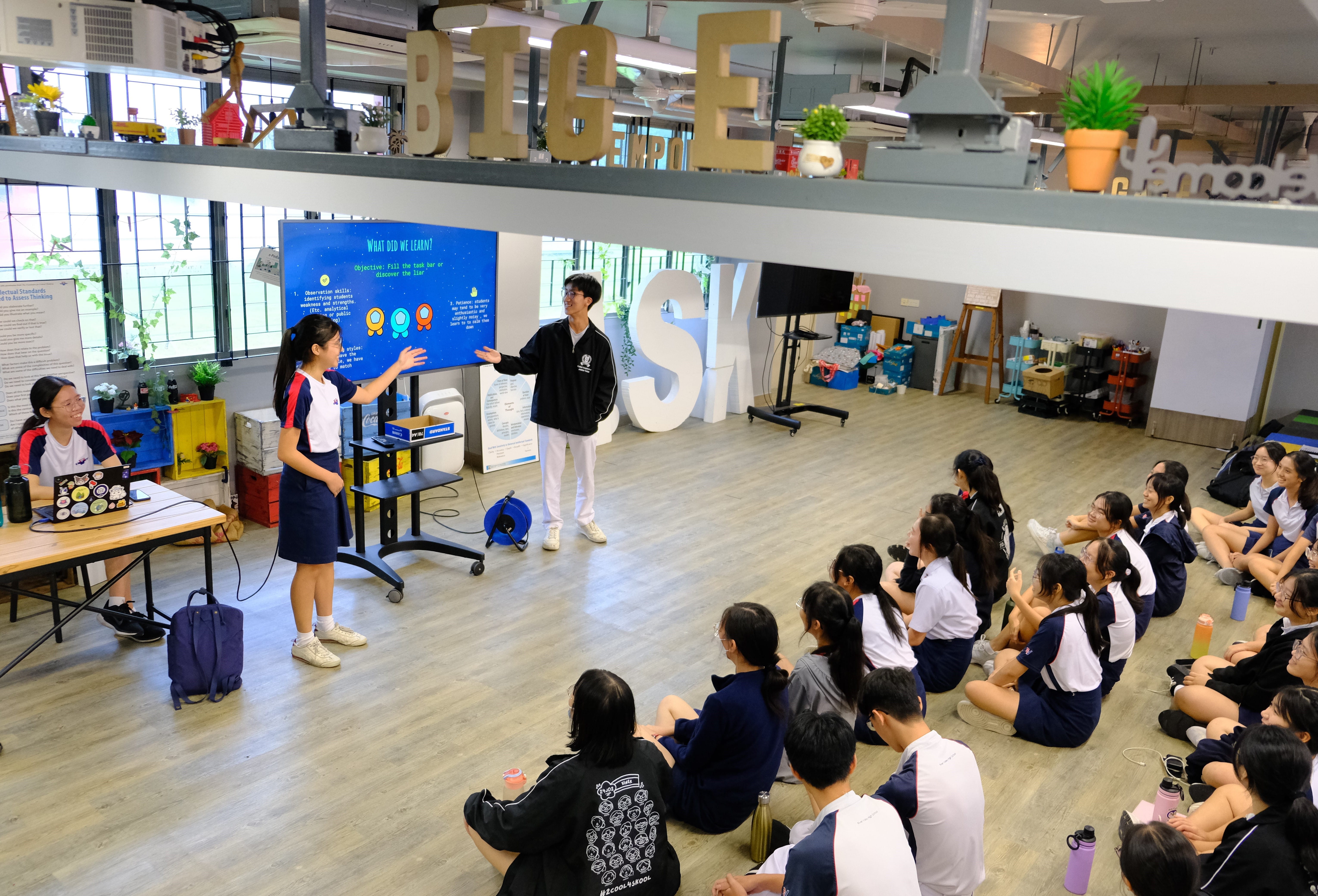 Students seated on floor watch two presenters beside a screen in a makerspace classroom.