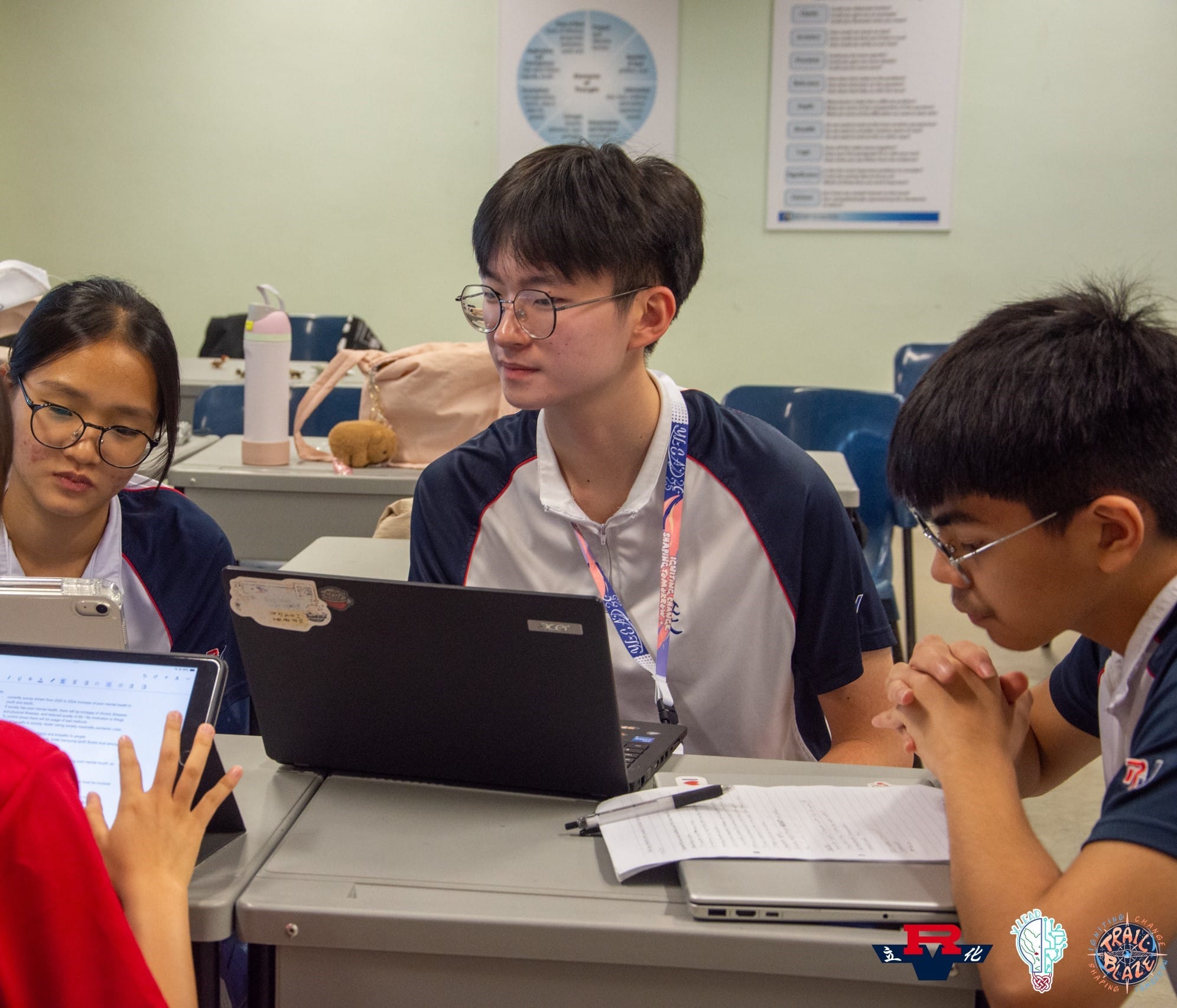 Three students in matching uniforms collaborate at a desk with a laptop, tablet, and papers in a classroom.