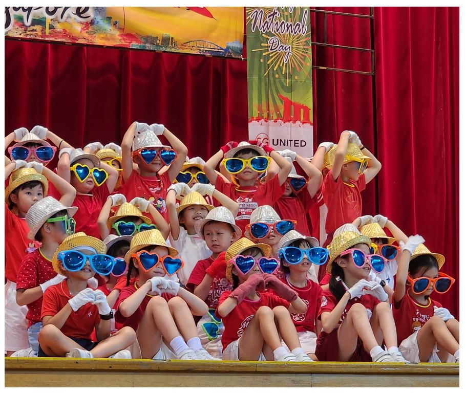 Children in red shirts, gold hats, and oversized colorful sunglasses pose on a stage at a National Day event.