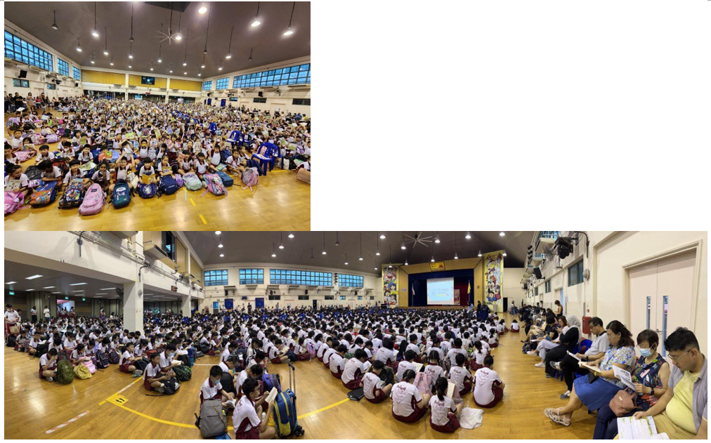 Large groups of students in uniforms sit on a school hall floor during an assembly, with adults seated nearby.