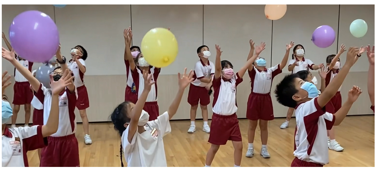 Children in school uniforms and face masks reach up to toss colorful balloons in an indoor hall.
