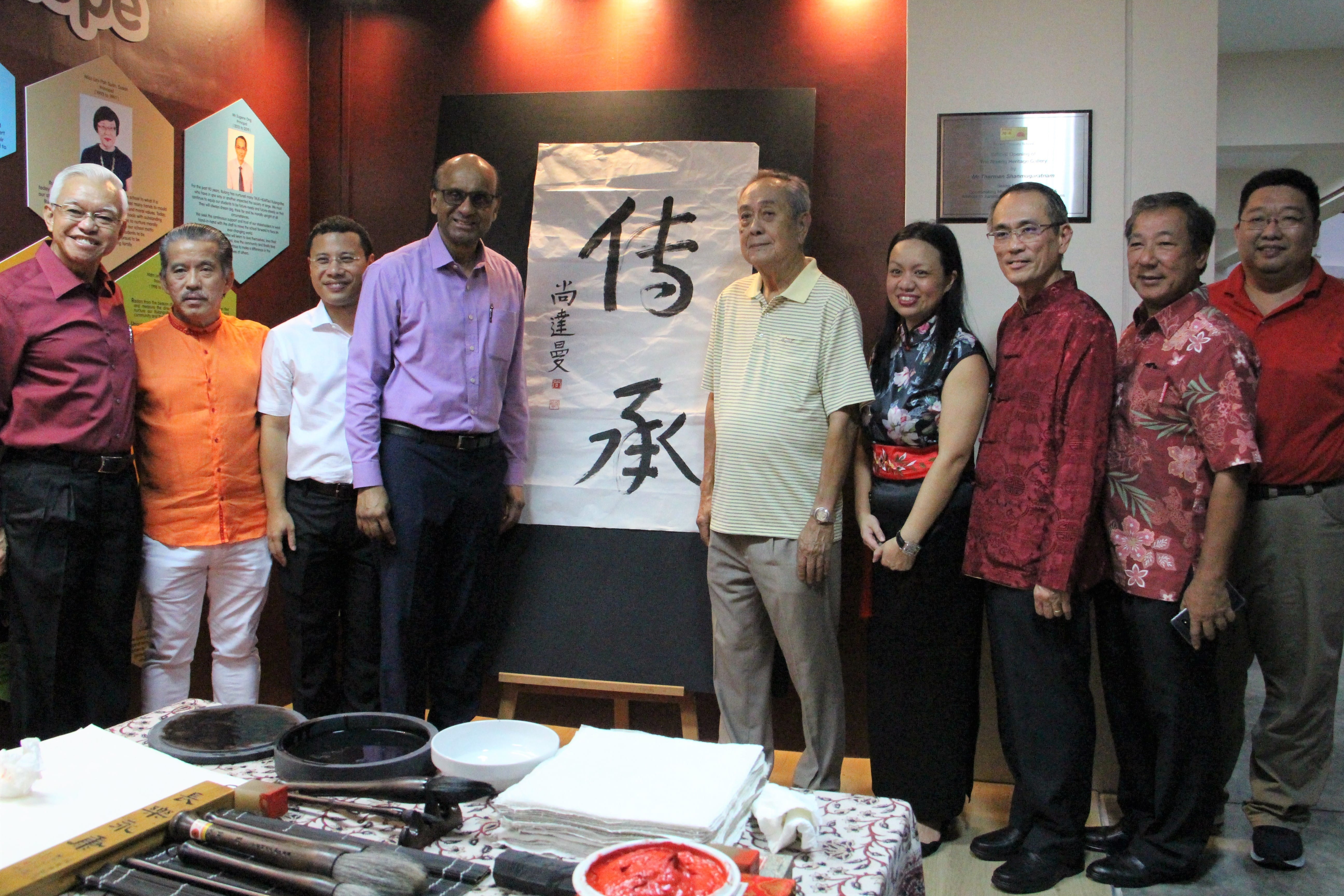 Nine people stand together smiling beside a large Chinese calligraphy scroll on an easel, with brushes and ink on a table in front.