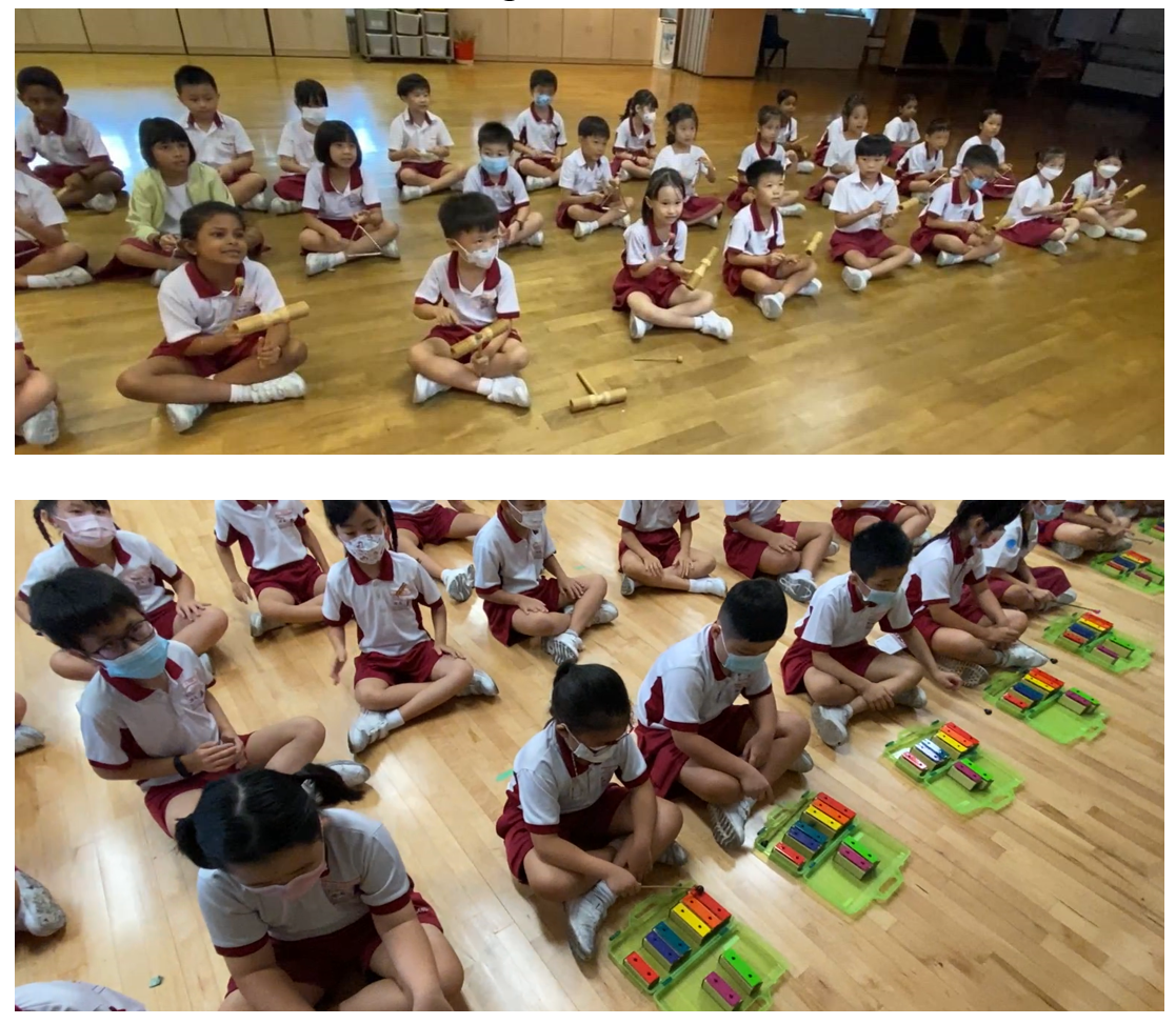 Young students in red and white uniforms sit on a wooden floor holding rhythm sticks and colorful xylophones.