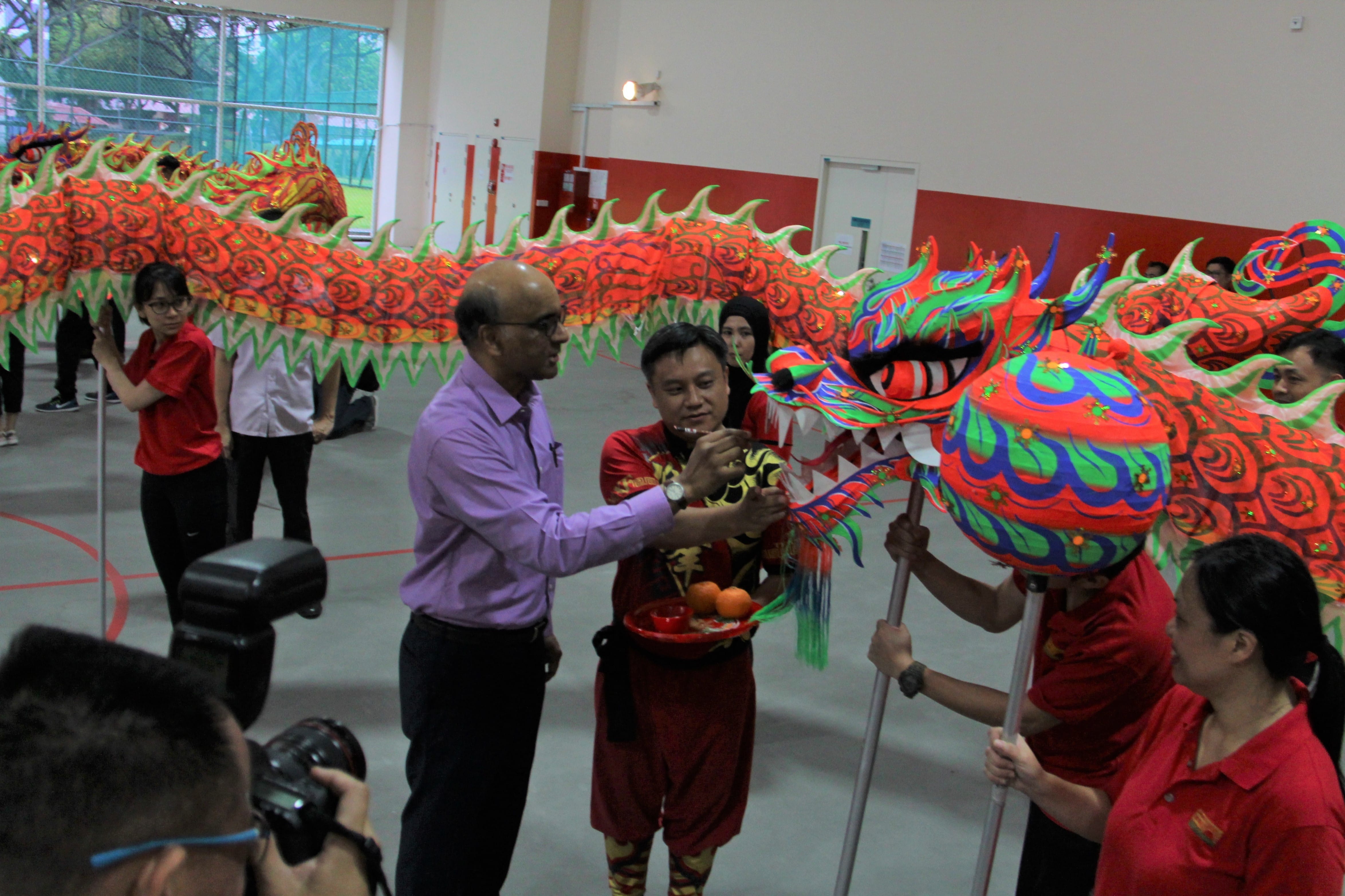 People perform a colorful red and green Chinese dragon dance indoors, with a man in purple shirt participating.