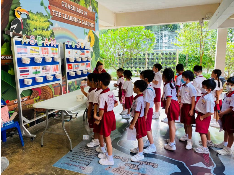 Young students in uniforms line up outdoors facing an ASEAN-themed learning board with flags displayed.