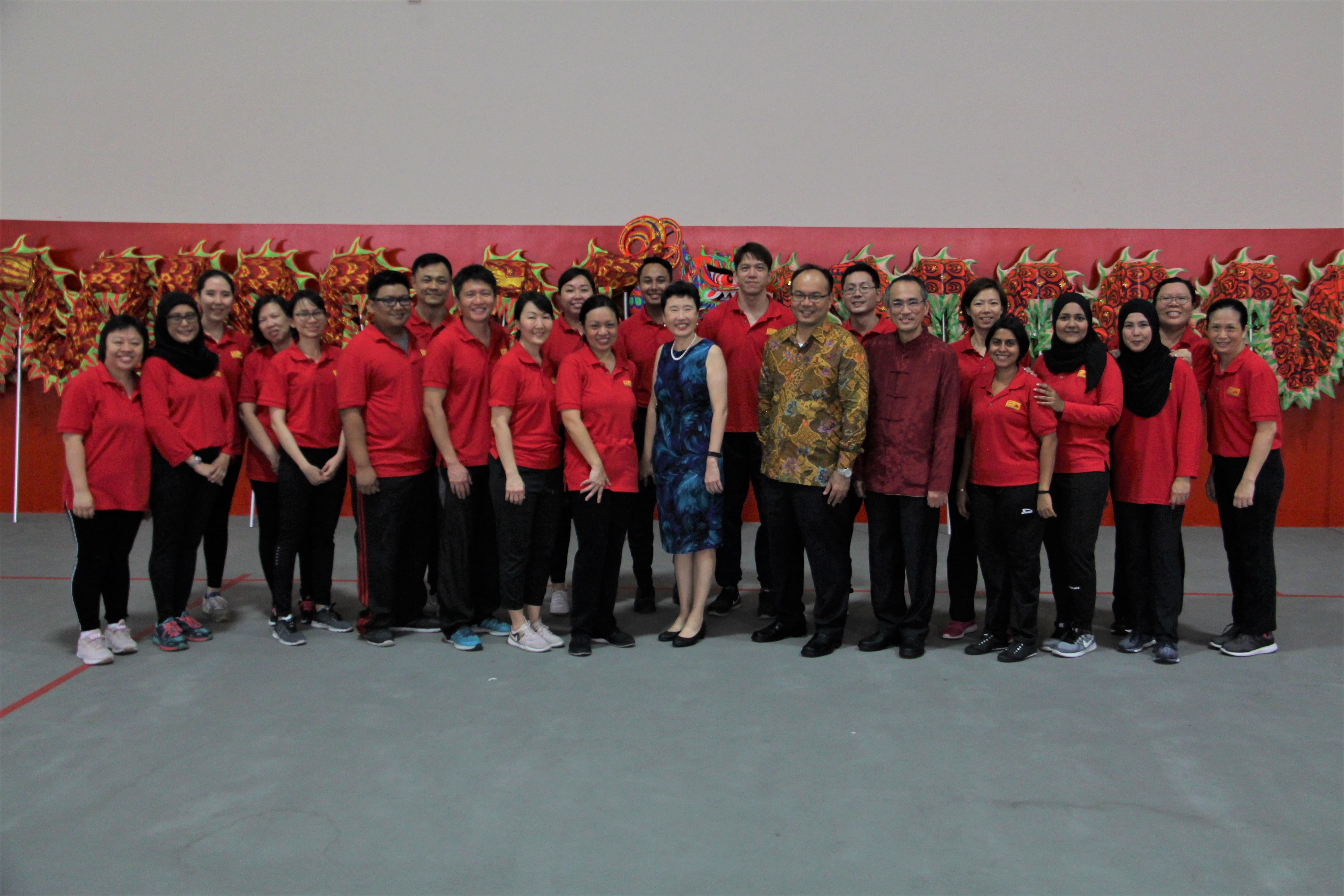 A large group of people in red shirts pose indoors in front of a colorful red dragon dance costume backdrop.