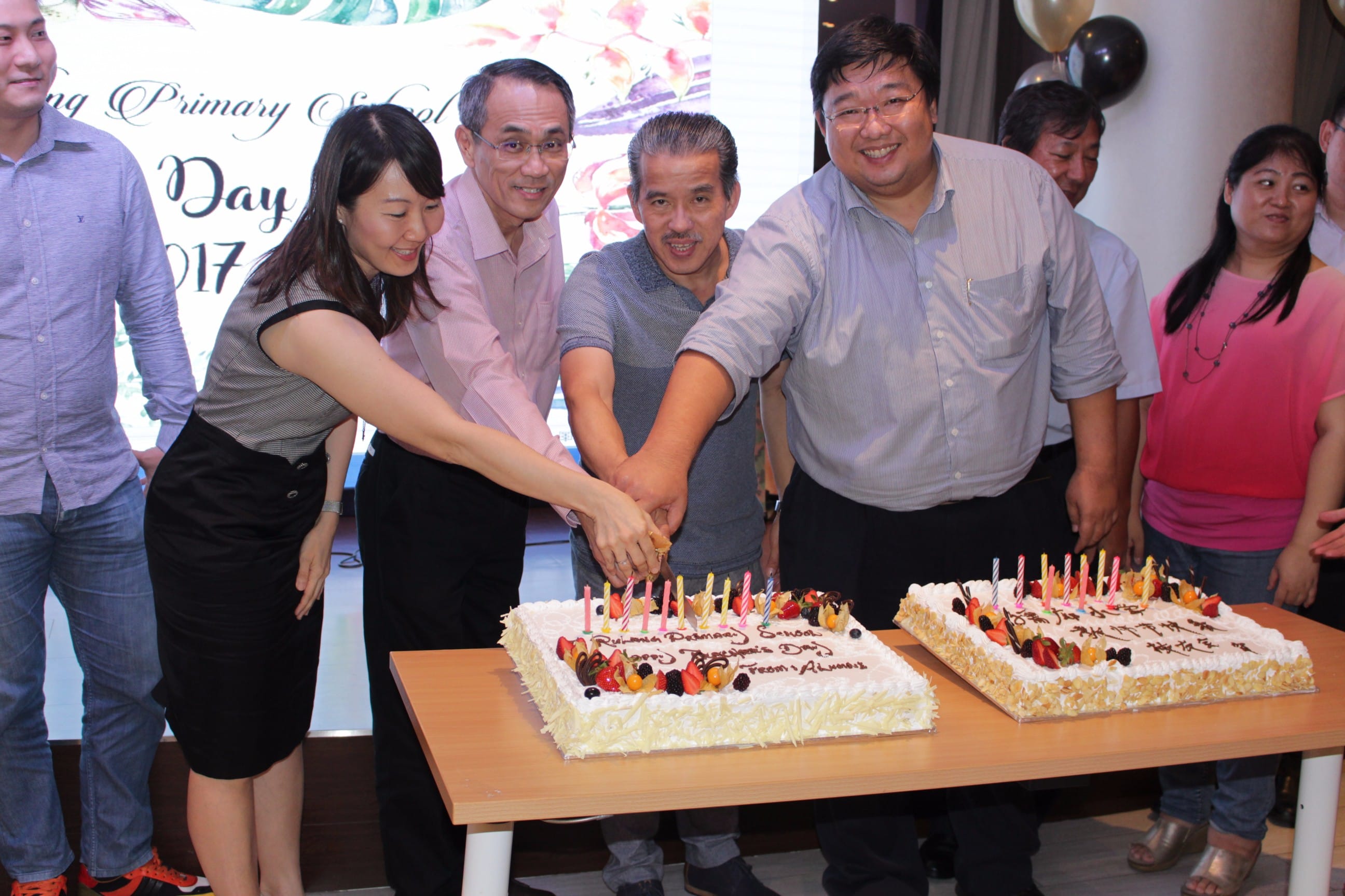 Four people cutting two decorated celebration cakes with candles at a primary school Teachers' Day event in 2017.