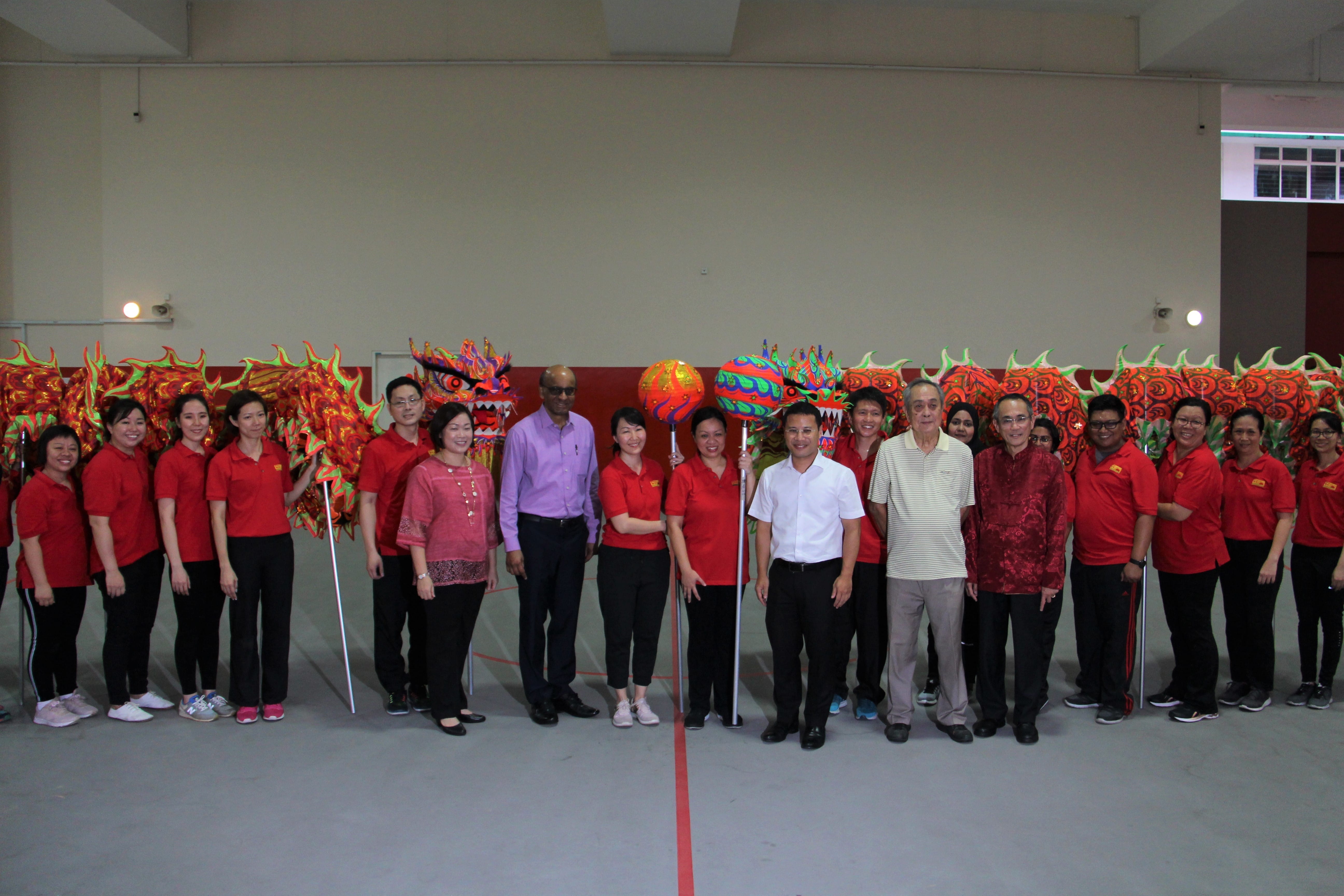 Group of people in red shirts posing indoors in front of colorful Chinese dragon dance props on poles.