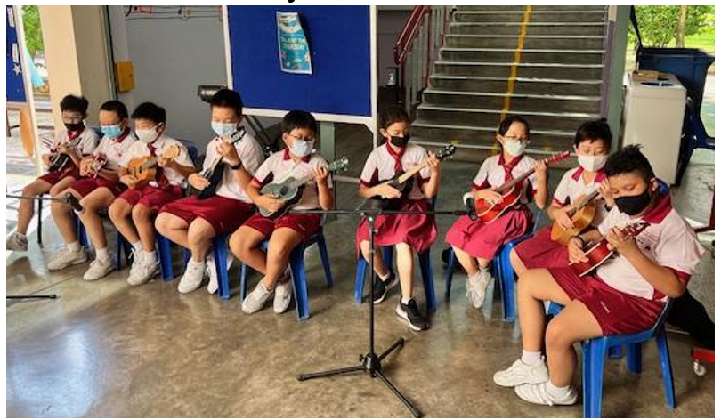 Eight young students in red uniforms and face masks sit on blue stools playing ukuleles indoors.