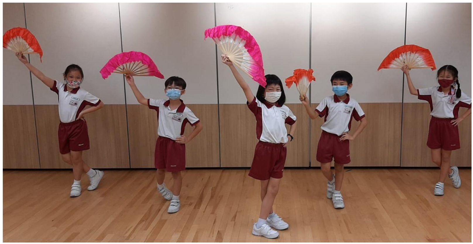 Five masked school children in uniforms pose while holding colorful pink and orange Chinese dance fans in a studio.