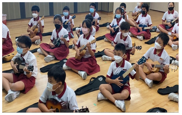 Young students in red and white uniforms and face masks sit on a wooden floor holding ukuleles during a music class.
