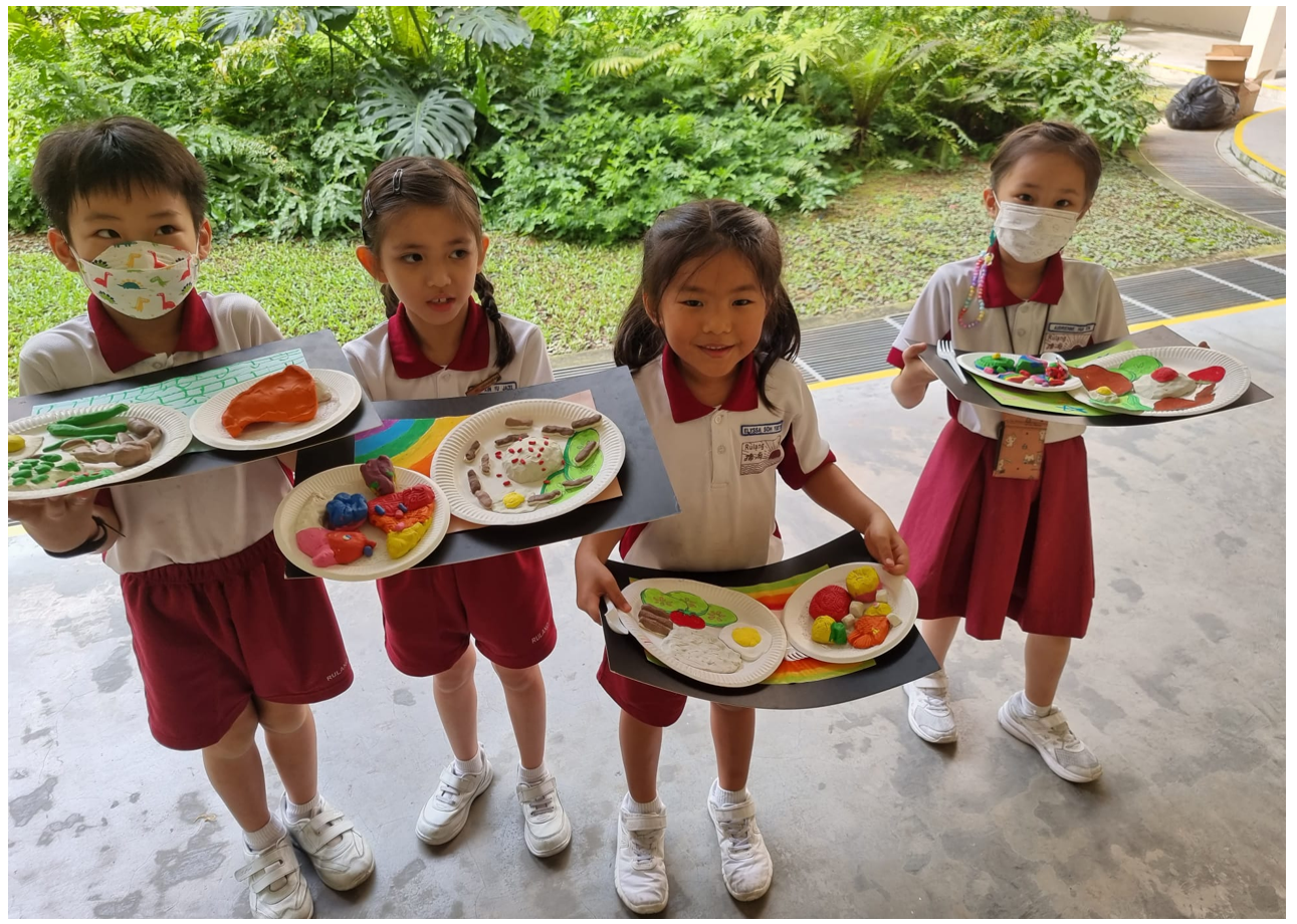 Four young students in red and white uniforms hold paper plates with colorful clay food models outdoors.