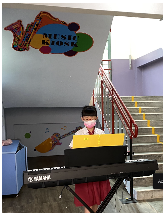 A young girl wearing a mask plays a Yamaha keyboard near a colorful Music Kiosk sign by a staircase.