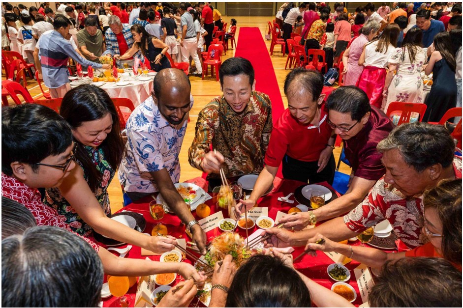A diverse group of smiling people toss a dish together with chopsticks at a large festive banquet with red decor.