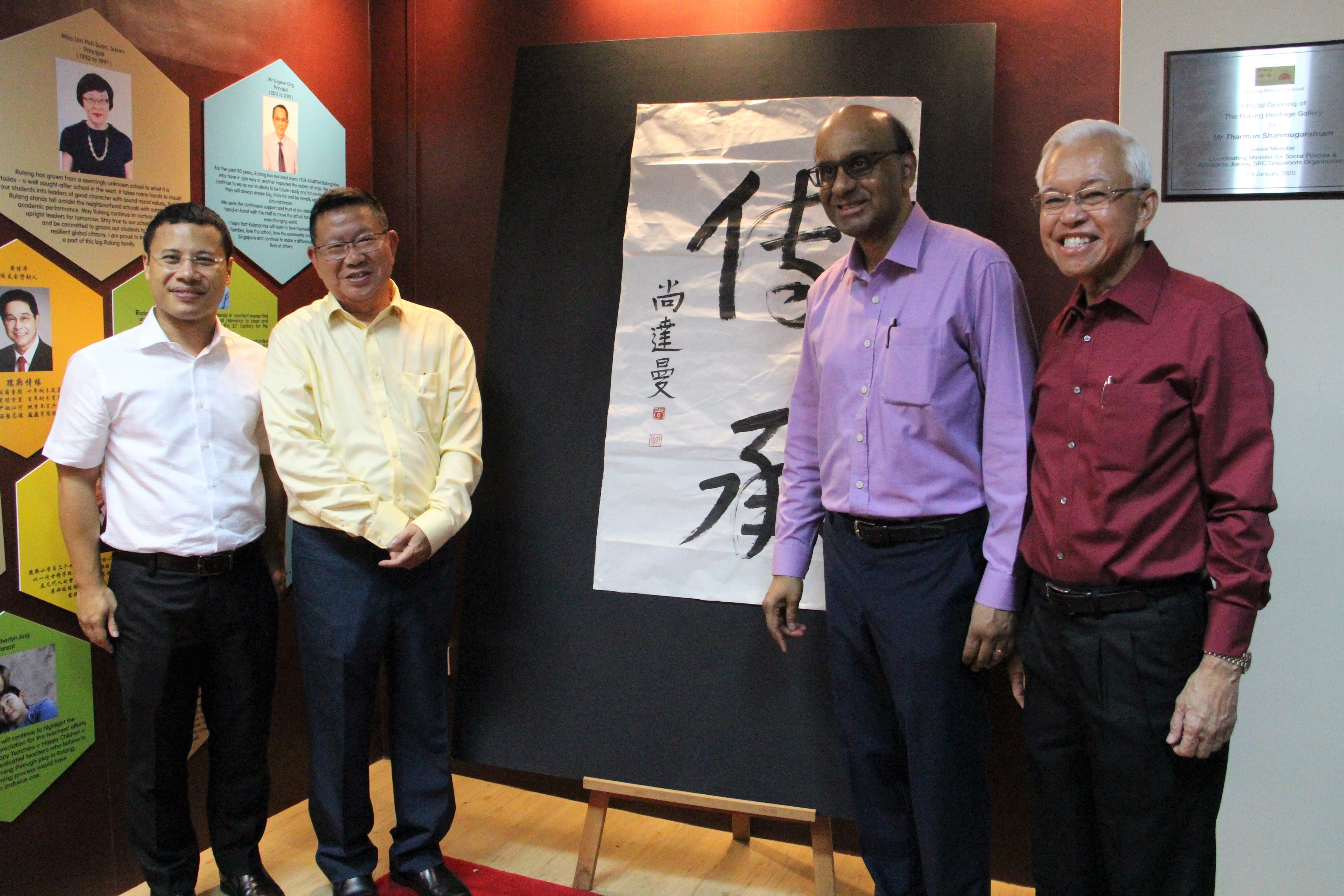 Four men smile while posing beside a large Chinese calligraphy artwork on an easel indoors.