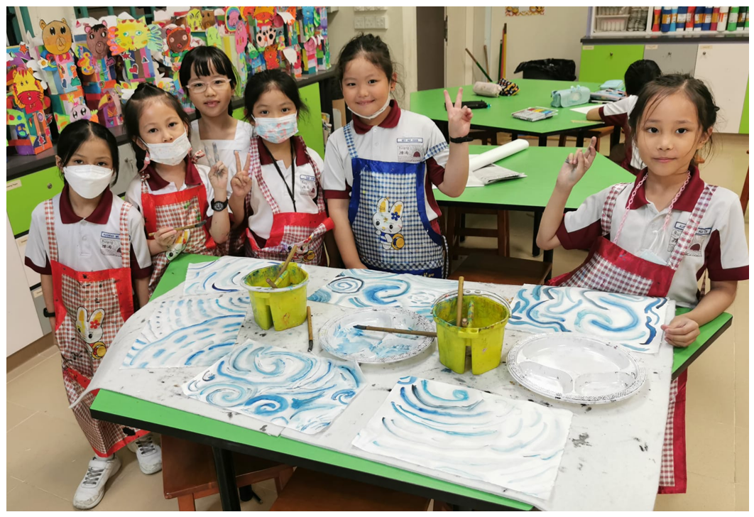 Six young girls in aprons pose with peace signs around a table covered in blue swirl paintings during an art class.