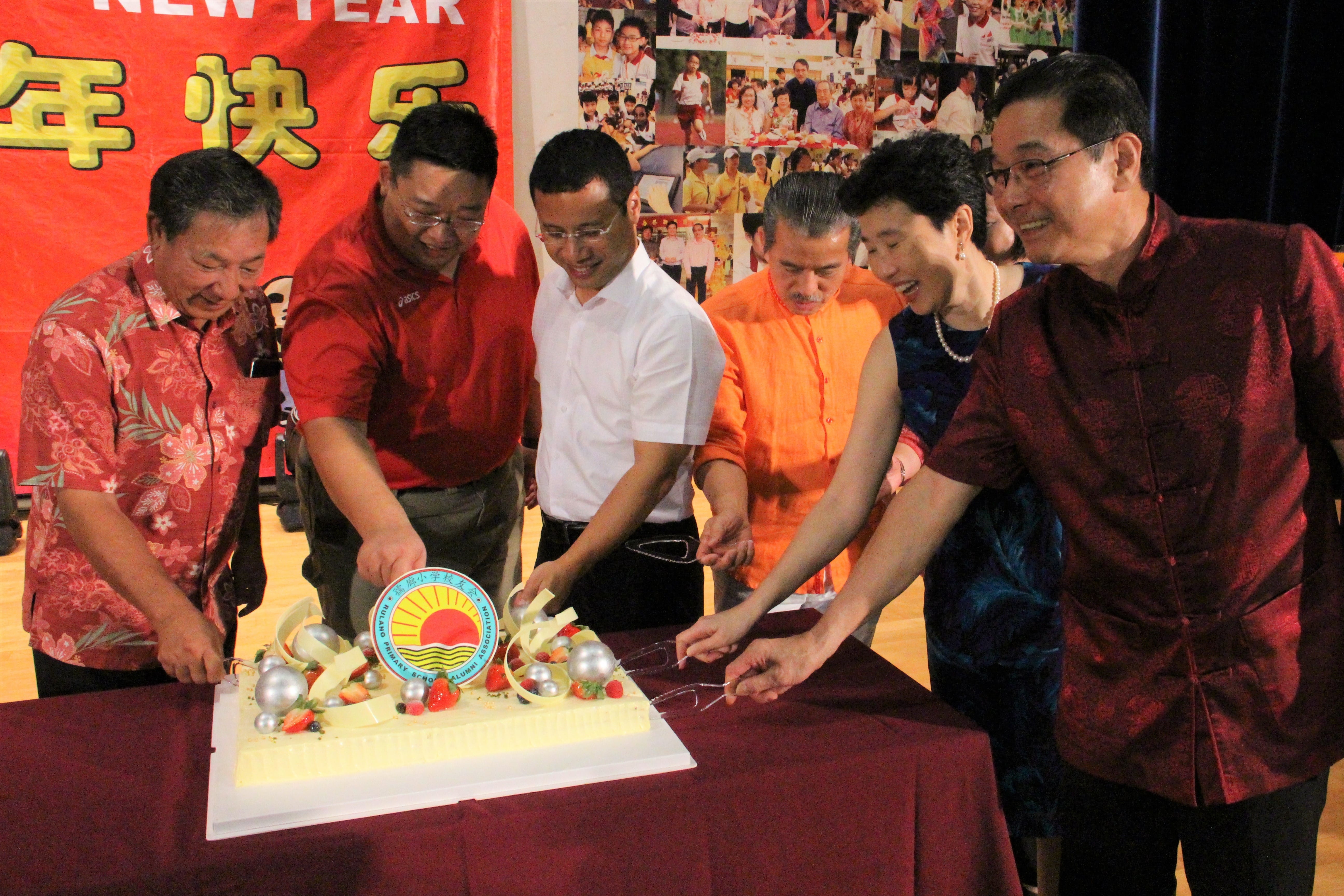Six people cutting a decorated cake at a Chinese New Year celebration with a red banner in the background.