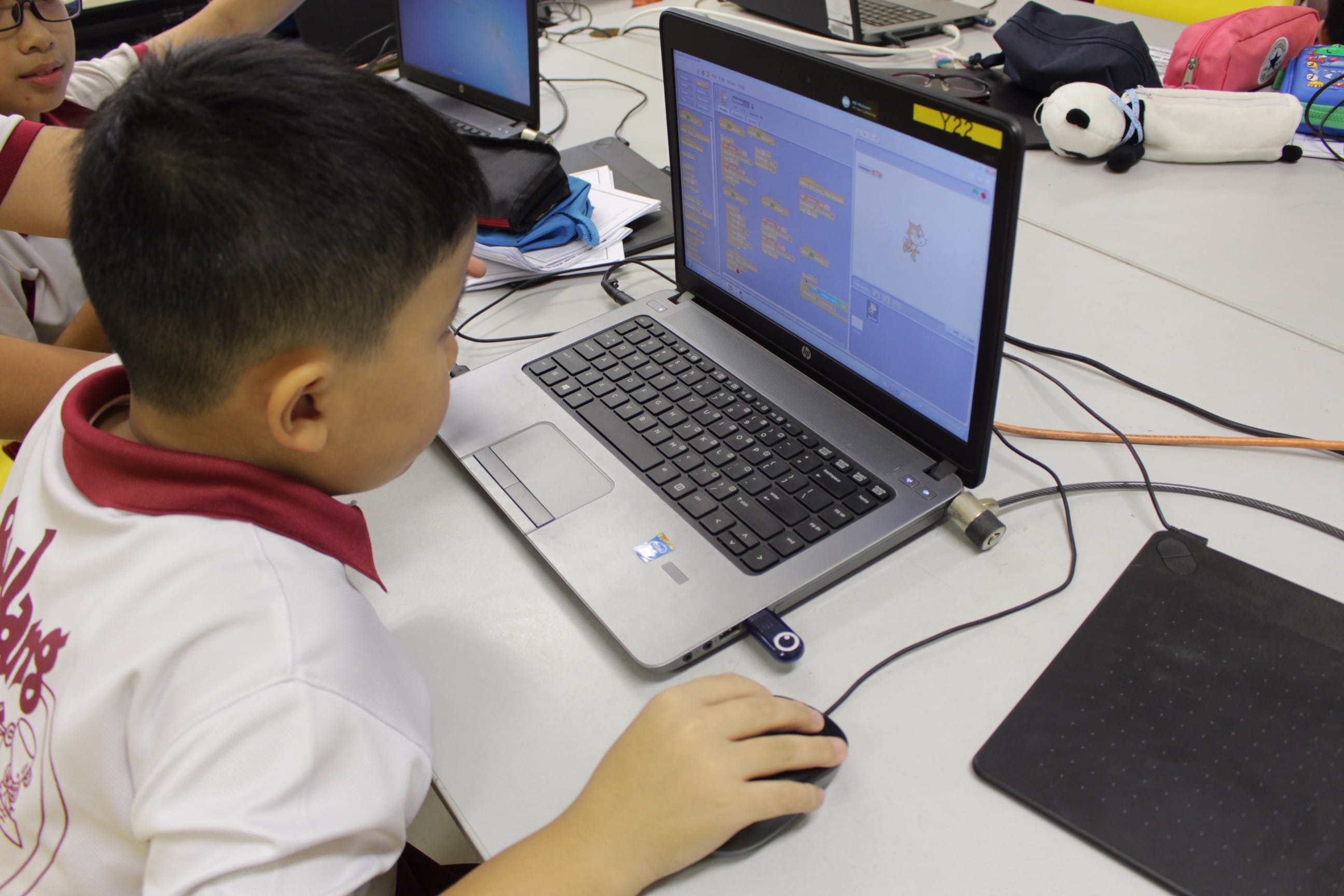 A young boy in a white shirt uses a mouse while working on a Scratch coding project on an HP laptop.