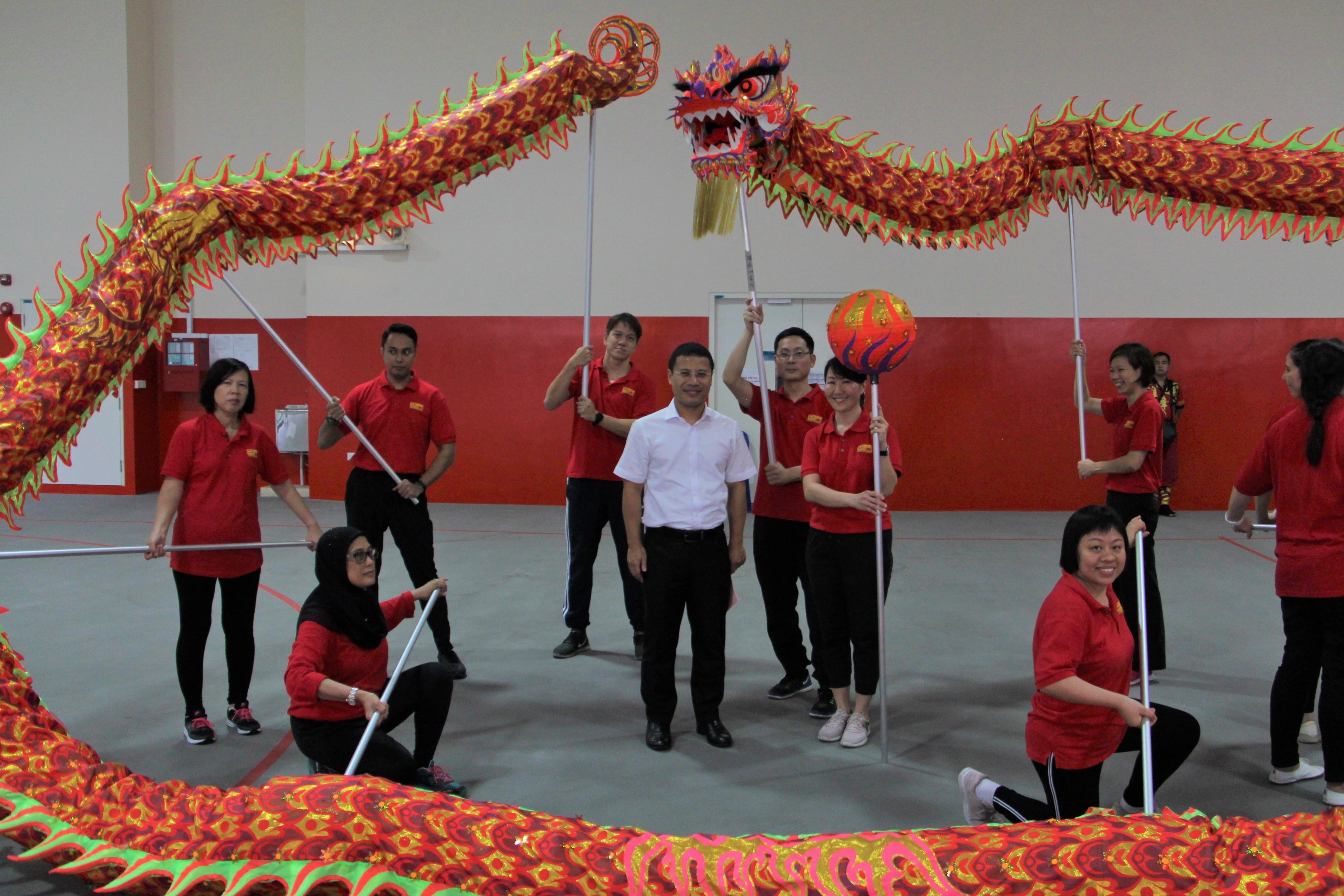 A group of people in red shirts perform a traditional Chinese dragon dance indoors, holding poles to control the colorful red dragon.
