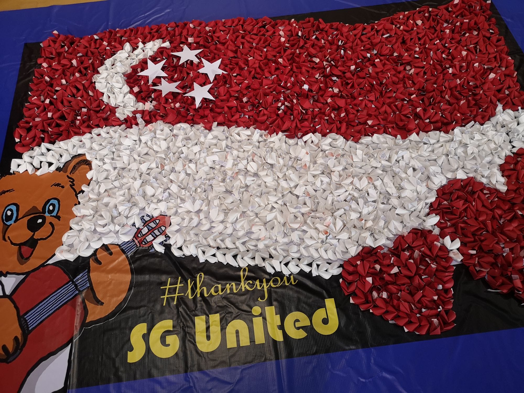 Singapore flag mosaic made of red and white origami pieces, with a bear mascot and text reading SG United.