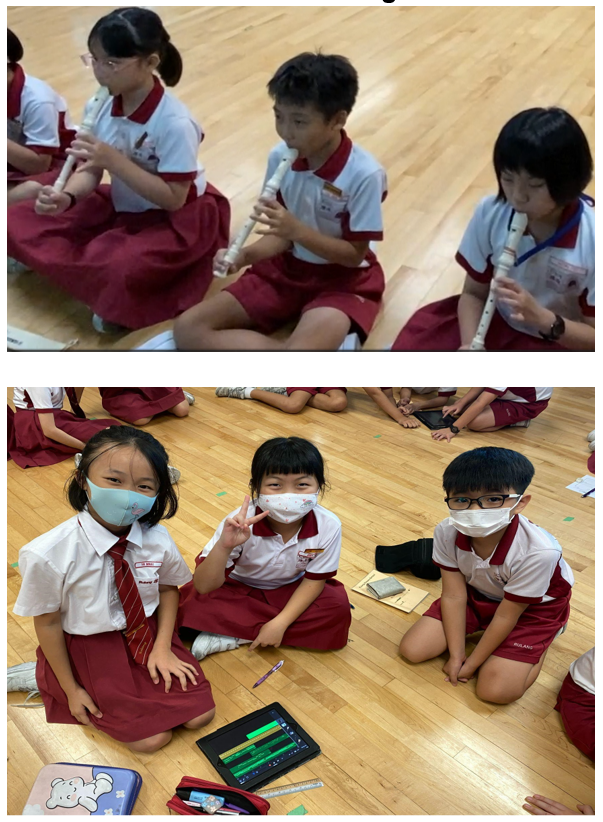 Young students in red and white uniforms sit on a floor, some playing recorders, others wearing masks near a tablet.