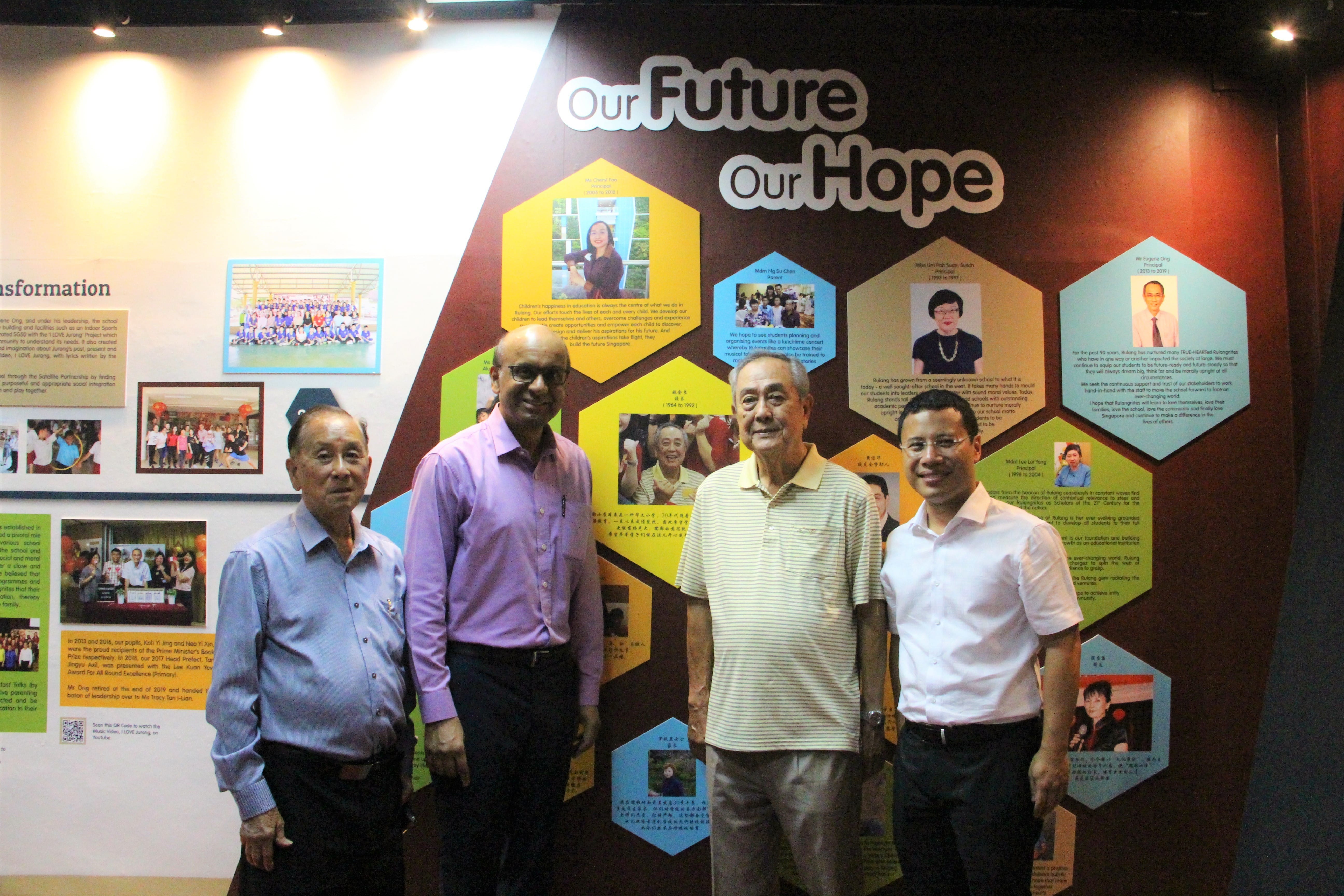 Four men stand smiling in front of a display wall reading Our Future Our Hope with hexagonal photo panels.