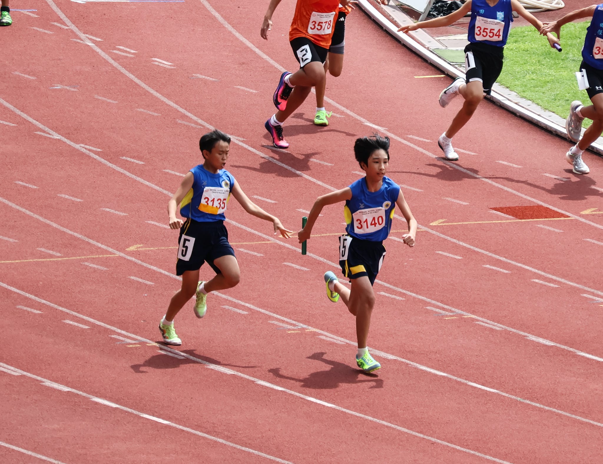 Two relay runners in blue completing a baton handover on the track, with other competitors visible running in adjacent lanes.