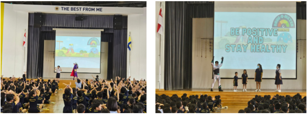 (left) Students seated on the hall floor raise their hands enthusiastically as a costumed superhero performer engages the crowd on stage, beneath a "THE BEST FROM ME" banner and a projected screen showing a colourful playground illustration. (right) The costumed performer stands with arms raised on stage next to two young students and two staff members, in front of a screen displaying "BE POSITIVE AND STAY HEALTHY" with a superhero-themed graphic, while the audience watches from the hall floor.