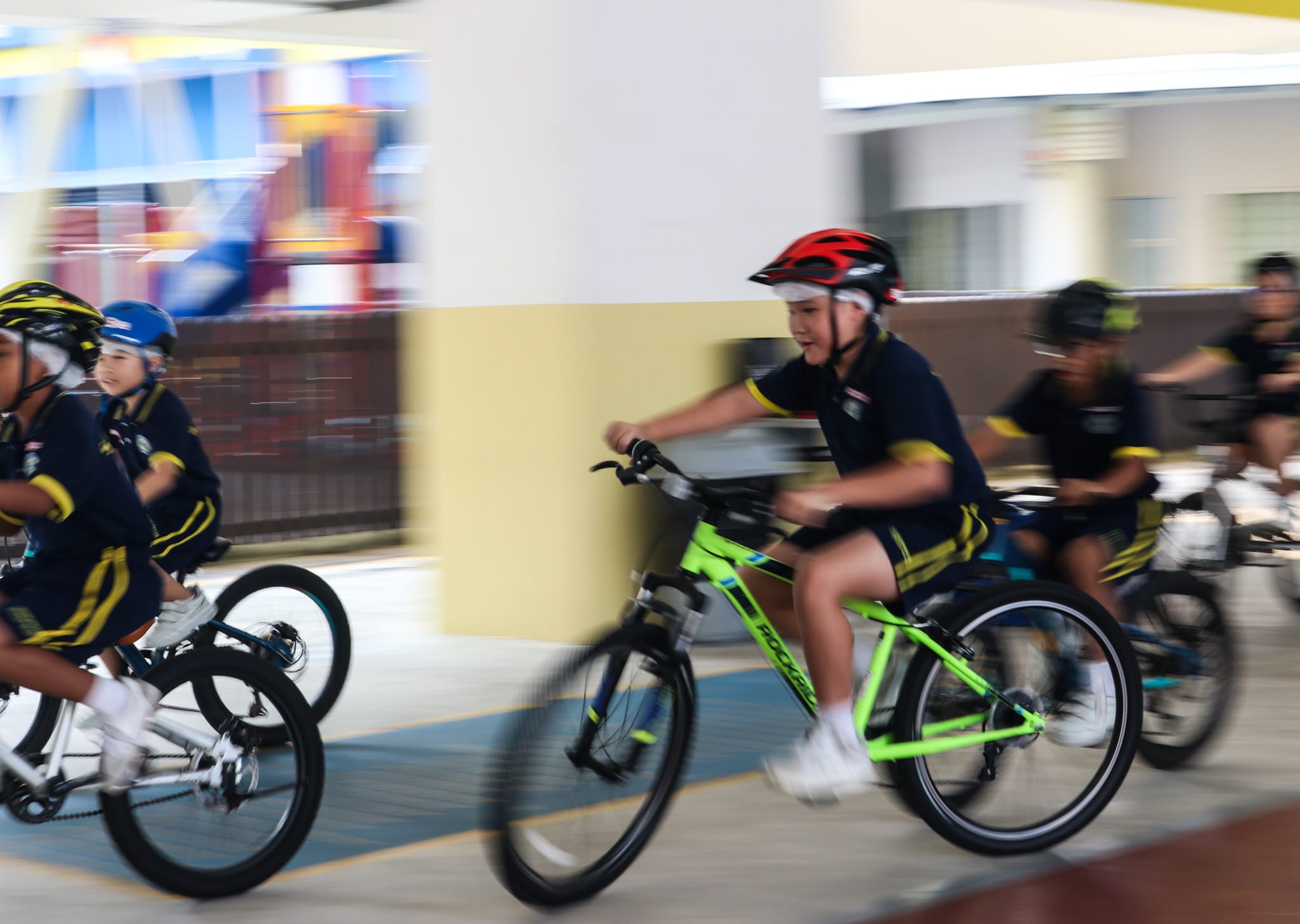 Several children in navy uniforms and helmets ride bicycles at speed through an indoor corridor.