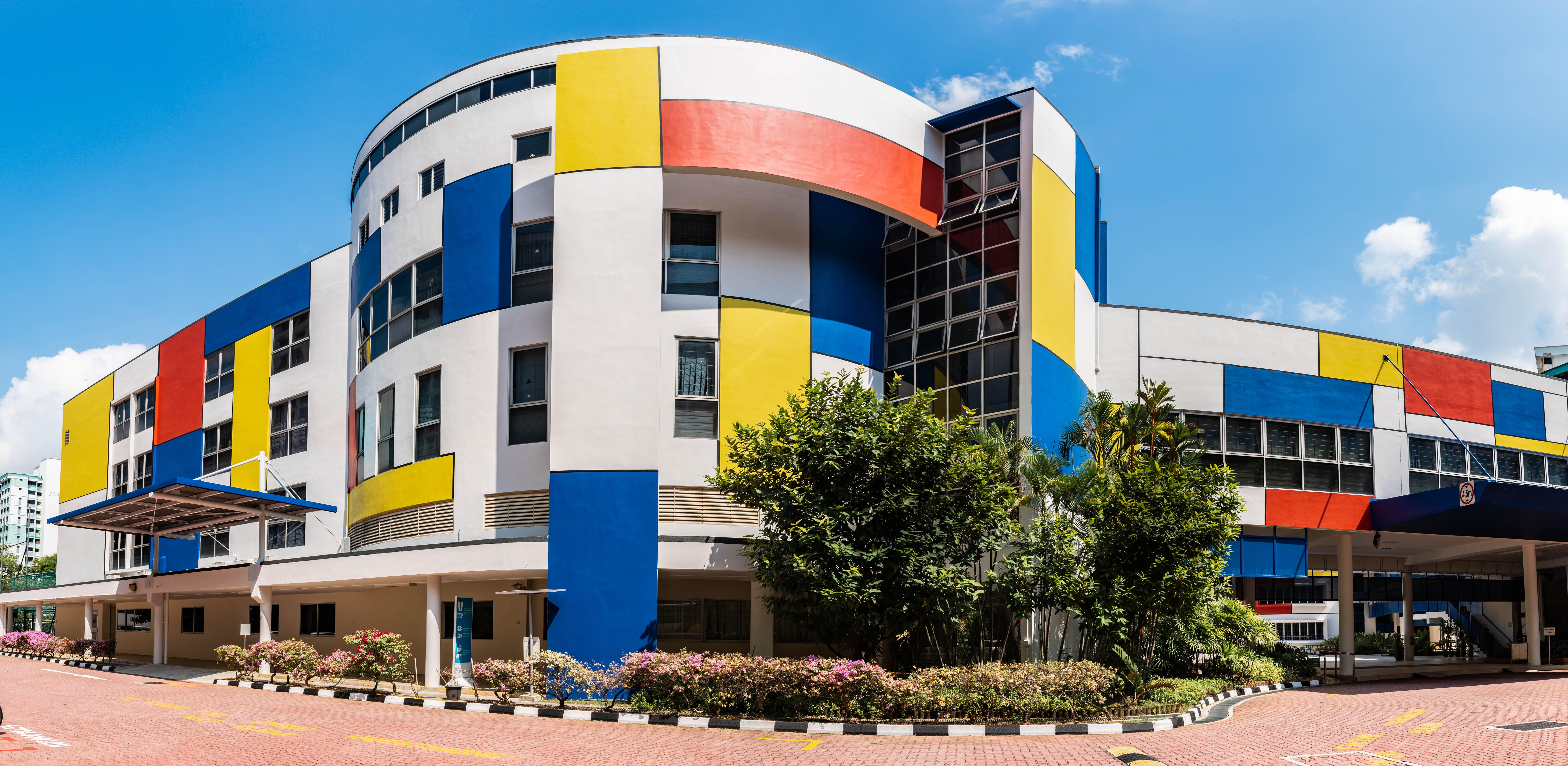 Curved white building with red, blue, and yellow panels, large windows, trees, and driveway under blue sky.