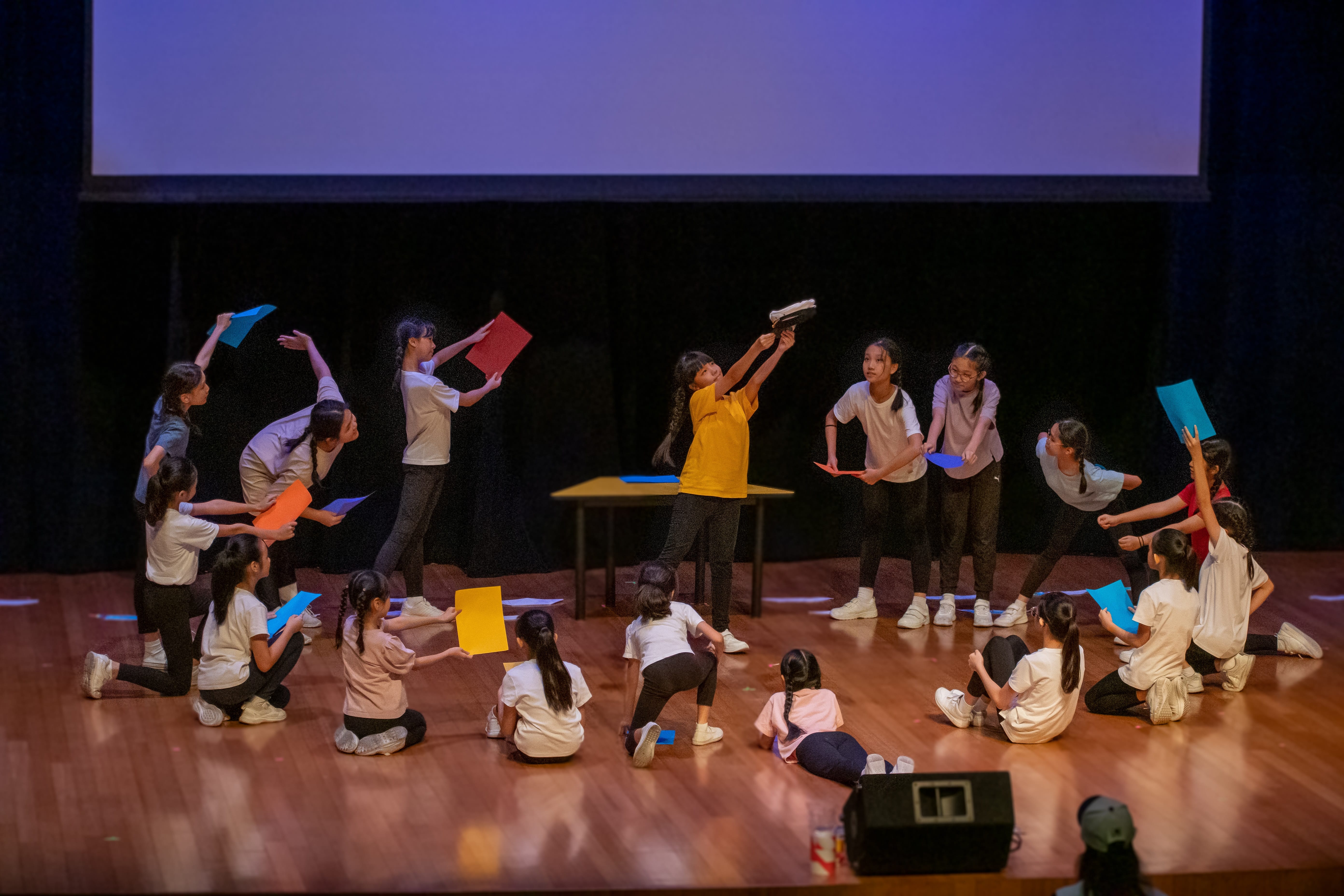 Group of children on stage holding colorful paper while one raises a shoe above a table under stage lights.