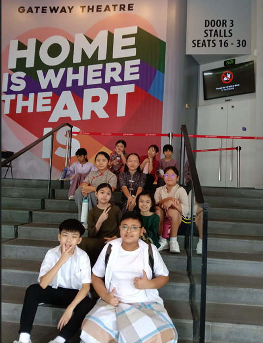 A group of students pose on steps at Gateway Theatre, in front of a large colourful backdrop reading "HOME IS WHERE THE ART IS," with signage for "Door 3, Stalls, Seats 16–30" visible nearby.