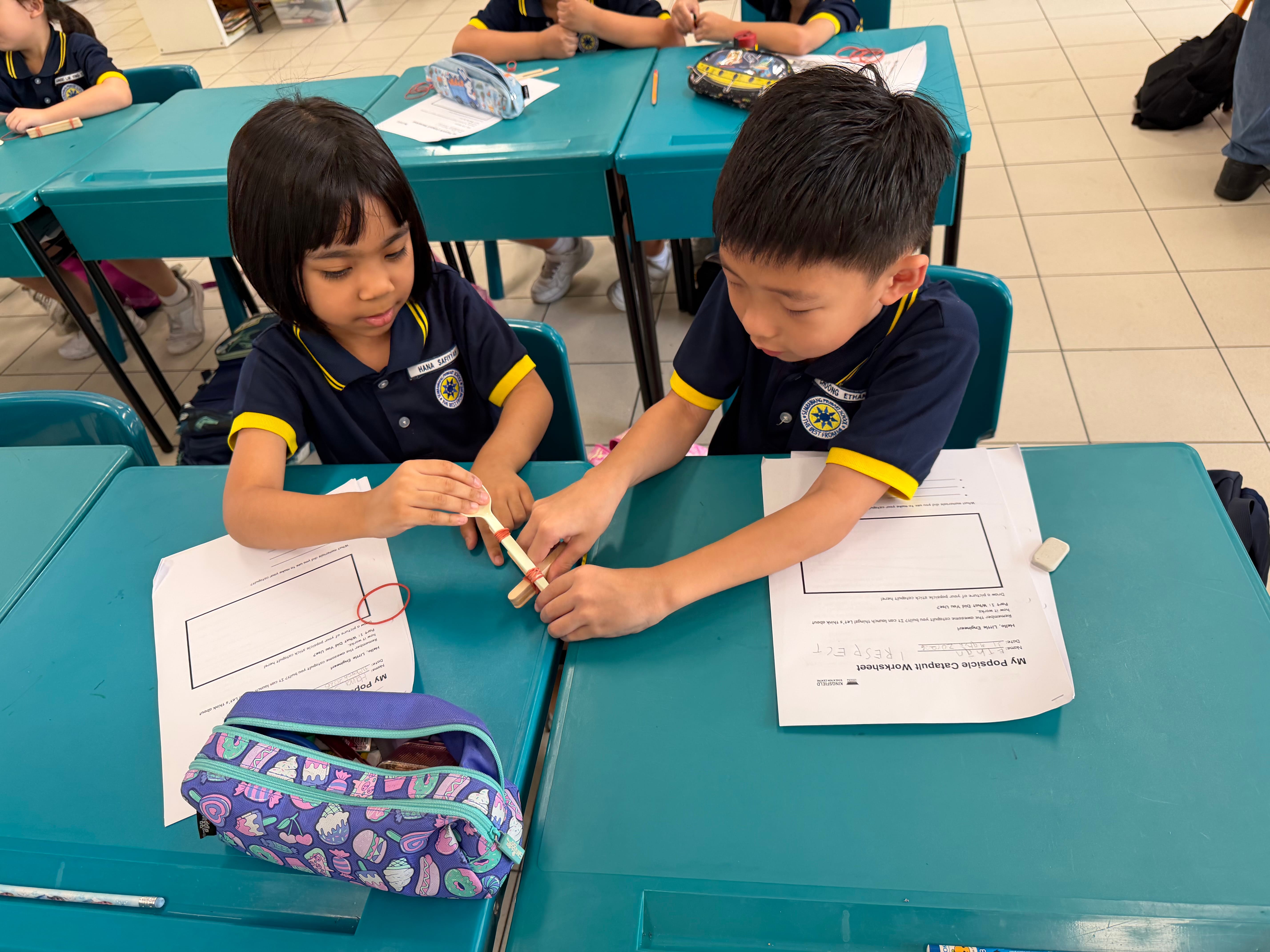 Two young students in navy uniforms build a popsicle stick catapult at a teal desk with worksheets nearby.