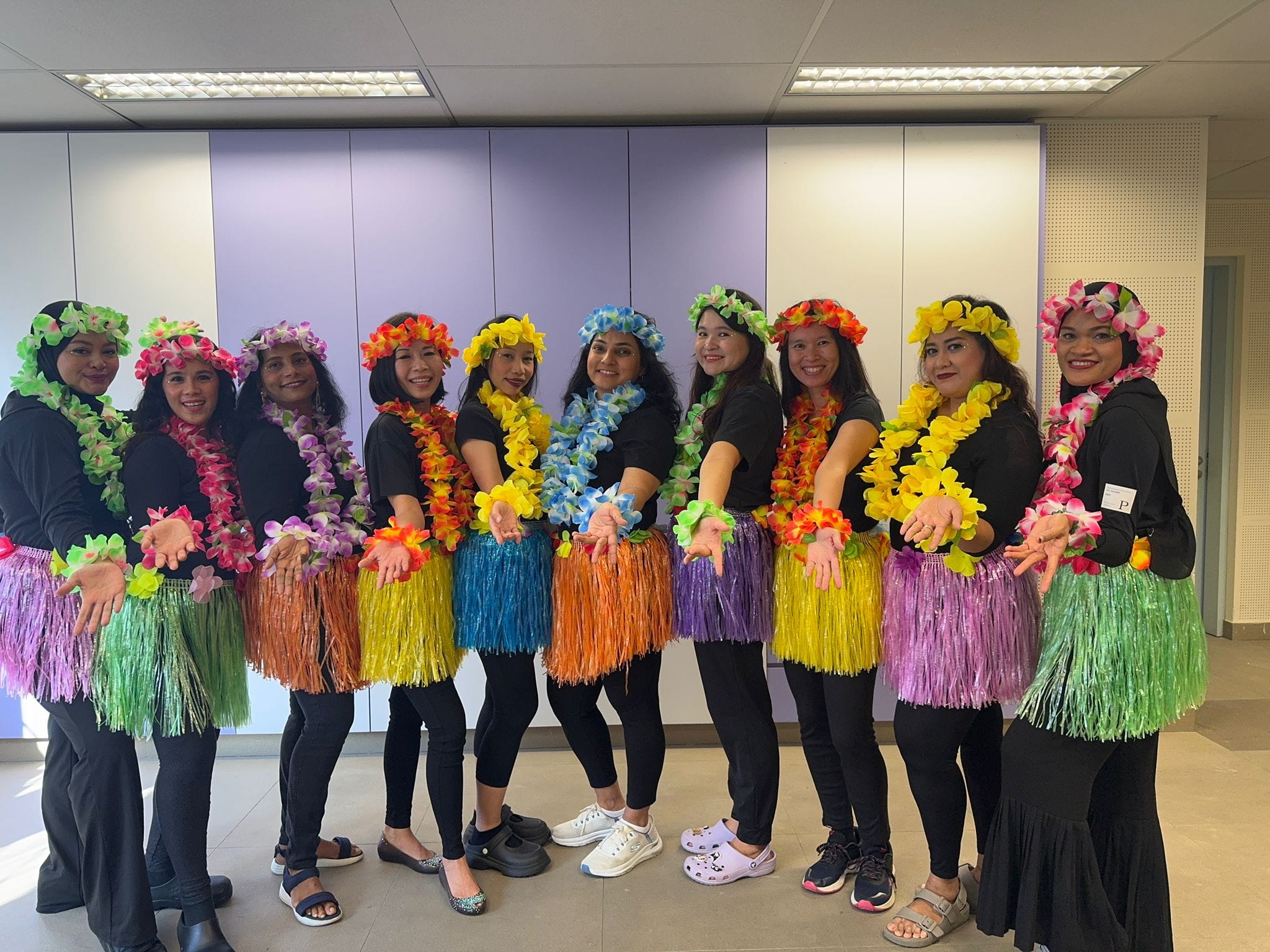 Ten women in black outfits wearing colorful hula skirts, flower leis, and floral headbands pose indoors.