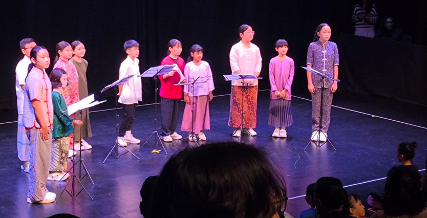 Students in traditional and cultural costumes stand on a dark stage at microphone stands with music stands in front of them, performing to an audience seated in the foreground.