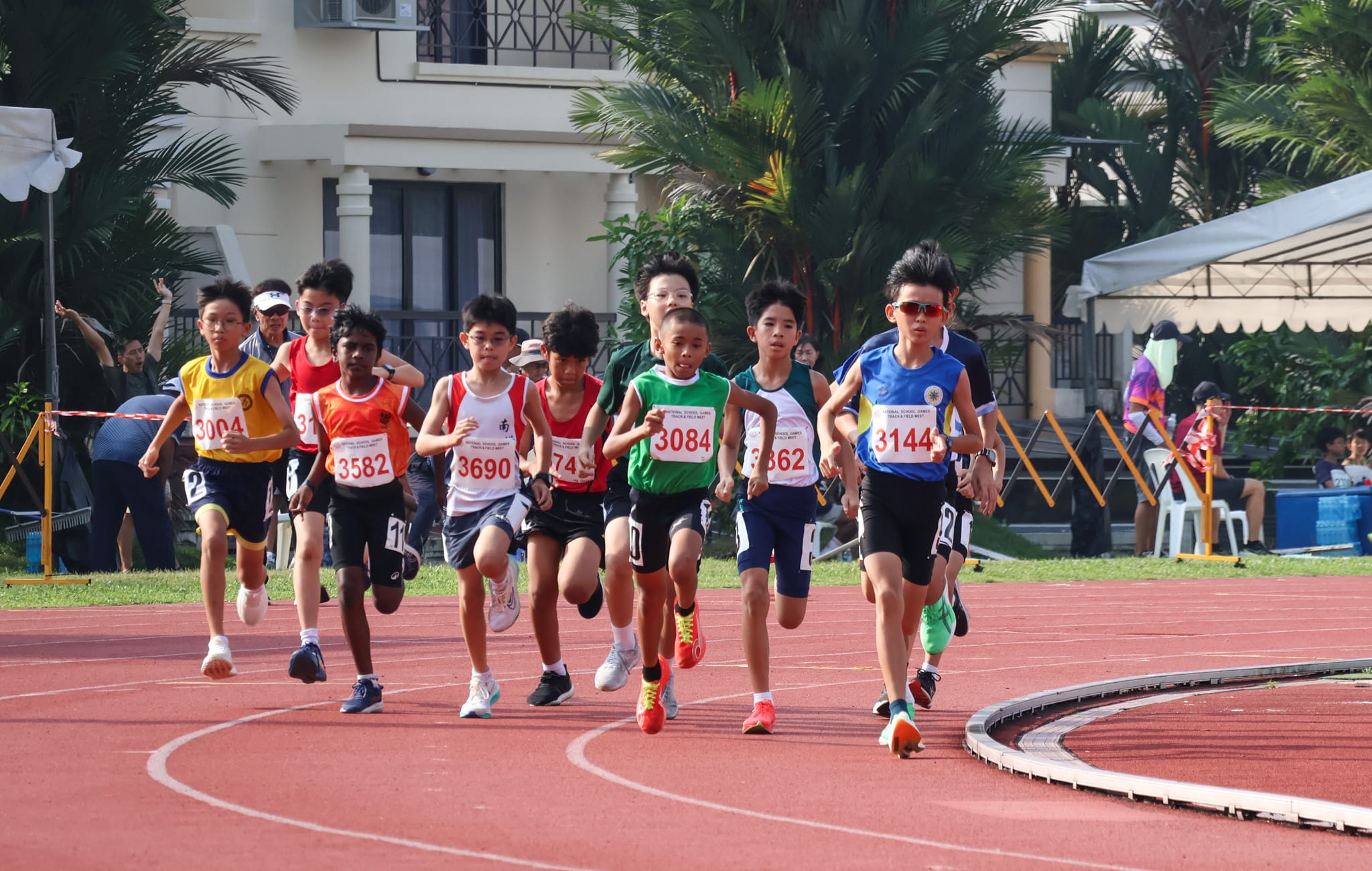 A pack of runners competing in a distance race at the National School Games Track and Field Meet, running along a red track past a grandstand and palm trees.